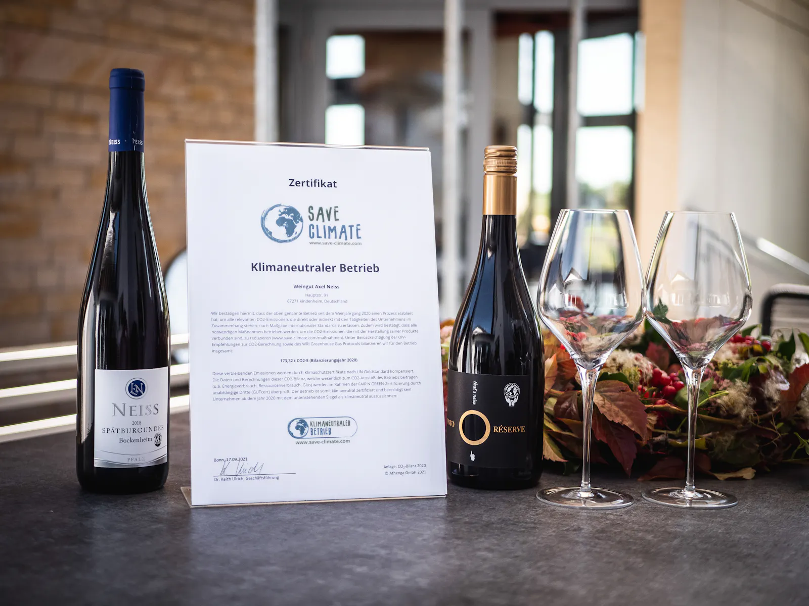 Das Weingut Neiss aus der Pfalz hat auf einem Tisch das Zertifikat "Klimaneutraler Betrieb" neben zwei Weinflaschen gestellt.
