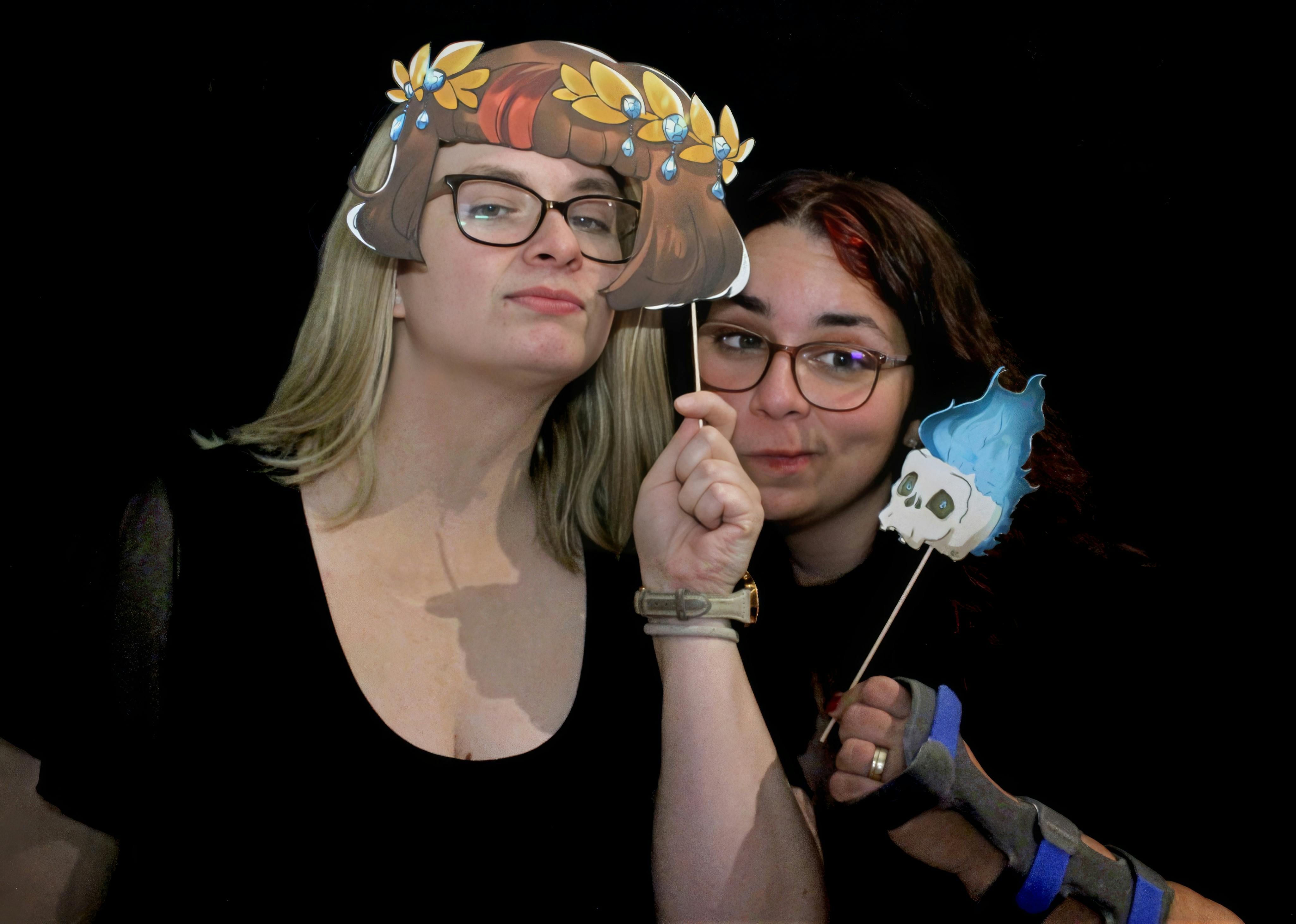 Links Carina, die eine Papierrequisite mit Perens-Karminoter-Schnitt Frisur an sich hält. Rechts daneben Aylin mit einem kleinen Papier-Gynther an einem Holzspieß, den sie neben sich hält. Beide posieren für ein Foto.