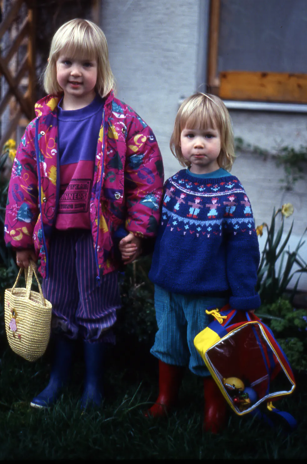 Altes Foto aus den 90ern: Zwei kleine blonde Mädchen stehen im Garten vor einem Haus, umgeben von Osterglocken. Die größere – Rieke – trägt eine bunte pink-gemusterte Jacke und hält einen geflochtenen Korb für die Ostereiersuche. Die kleinere – Randi – trägt einen blauen Norwegerpullover, rote Gummistiefel und schleppt eine bunte Kindertasche.