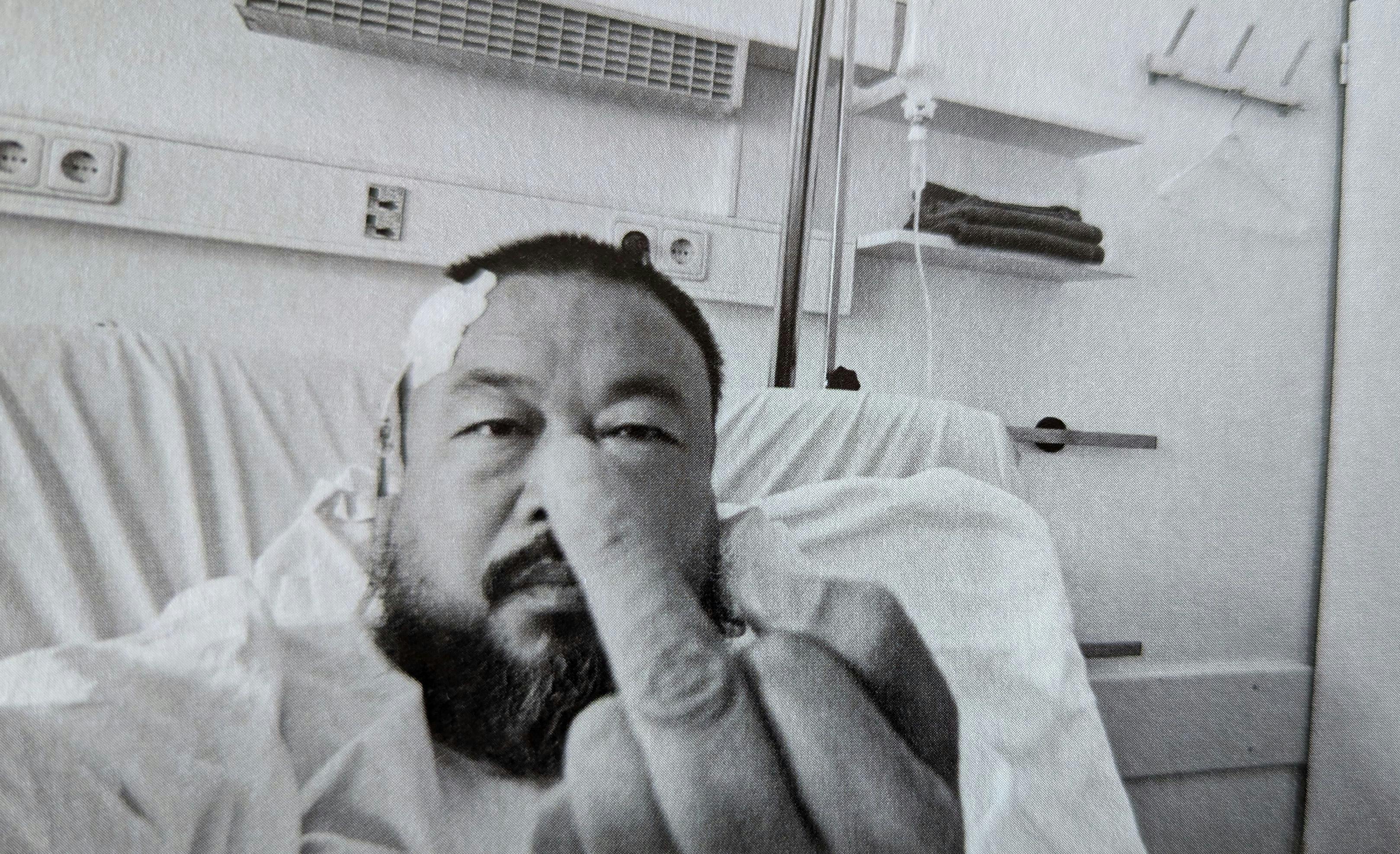 Der chinesische Künstler Ai Weiwei sitzt in einem Krankenhausbett. Sein Kopf ist verpflastert. Er hält einen Mittelfinger in die Kamera.