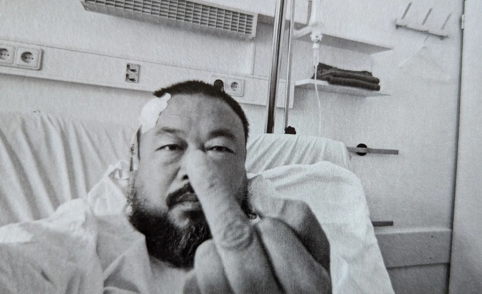 Der chinesische Künstler Ai Weiwei sitzt in einem Krankenhausbett. Sein Kopf ist verpflastert. Er hält einen Mittelfinger in die Kamera.