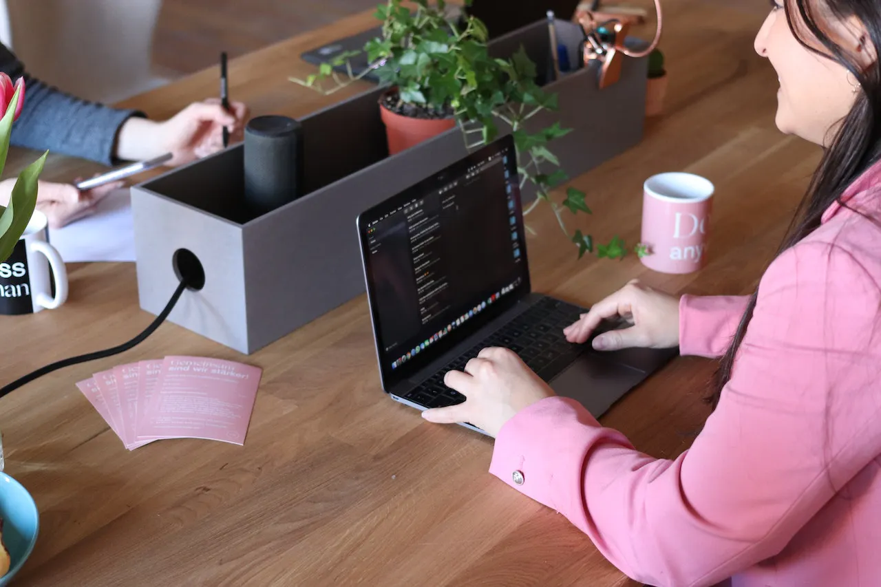 Frau in rosa Blazer vor einem Laptop an einem Tisch mit anderen Menschen.