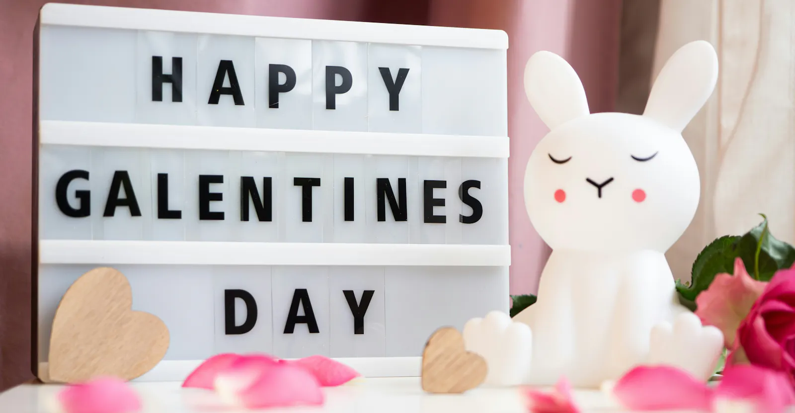 Auf einer Buchstaben-Tafel steht "Happy Galentines Day", daneben sitzt ein weißer Plastikhase mit roten Bäckchen und geschlossenen Augen. Um Tafel und Hase liegen Rosenblätter und eine Blüte