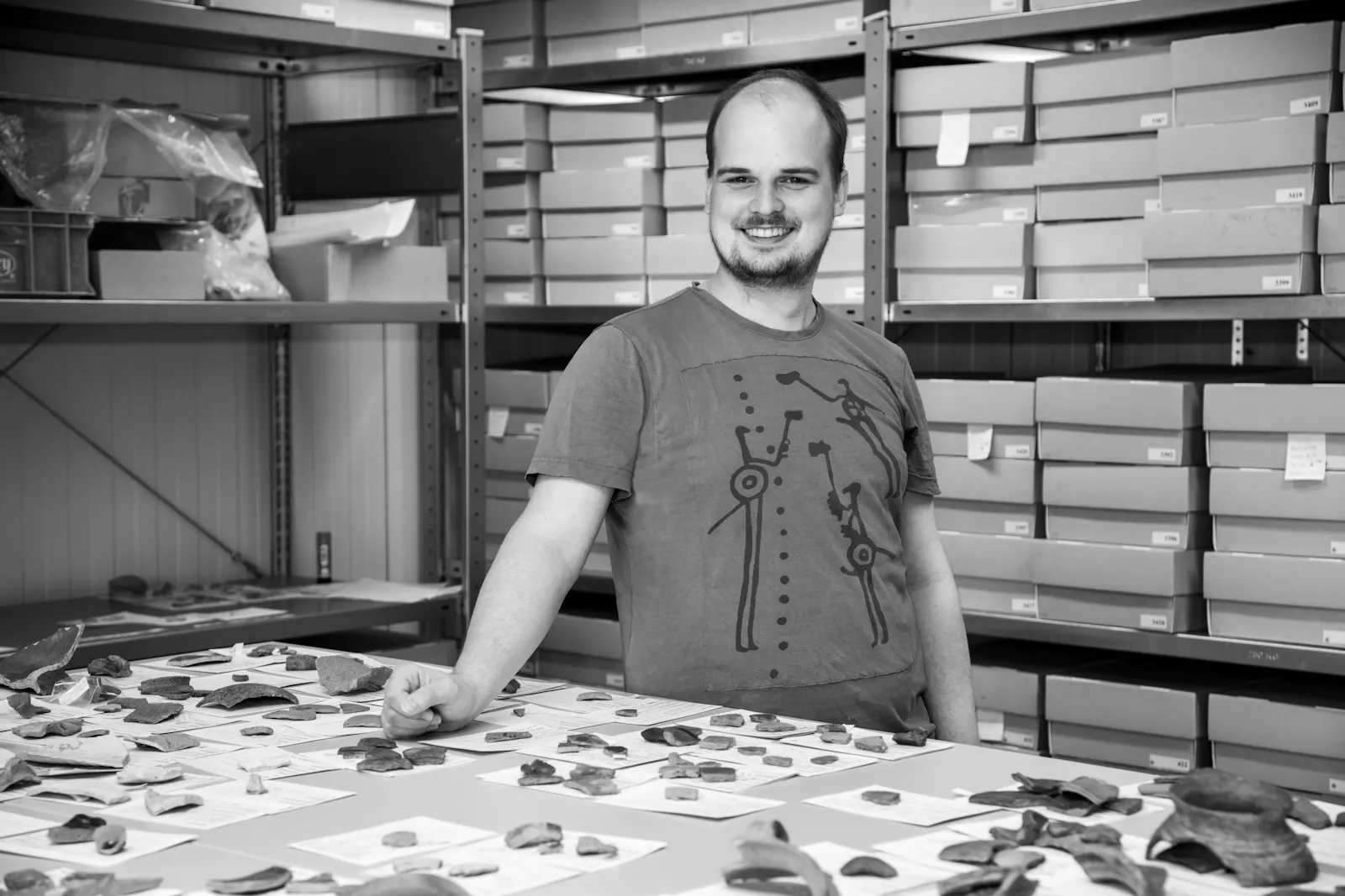 Johannes Reller steht hinter einem Tisch, auf dem Keramikscherben ausgelegt sind. Hinter ihm steht eine Regalwand mit Fundkisten.