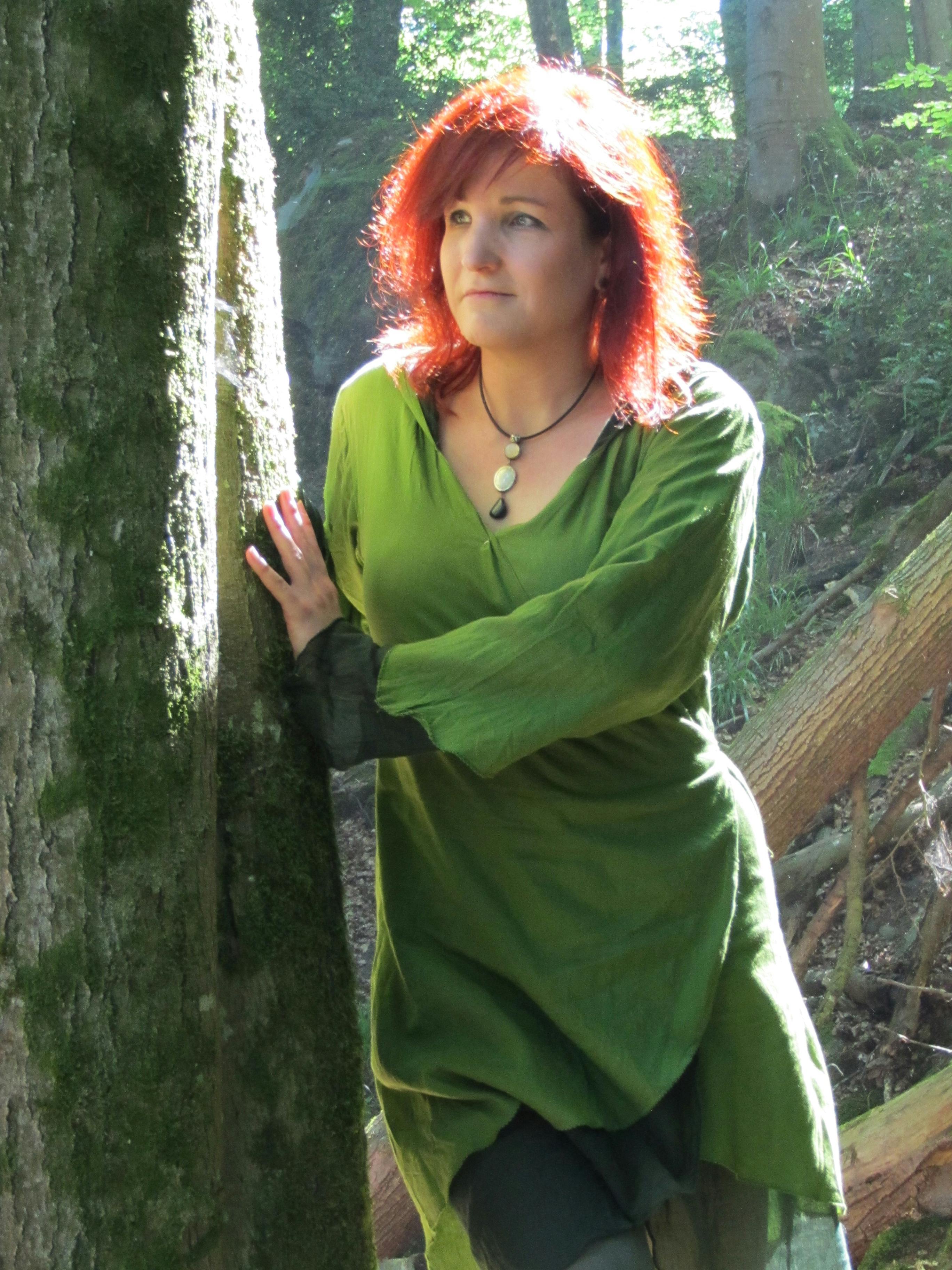 Foto von Sina Lys: Eine Frau mit leuchtend rotem Haar steht in einem Wald und lehnt sich an einen moosbewachsenen Baumstamm. Sie trägt ein olivgrünes Gewand mit weiten Ärmeln und eine schwarze Kette mit Steinanhängern. Ihr Blick ist nachdenklich in die Ferne gerichtet, das Sonnenlicht fällt durch die Baumkronen auf sie.