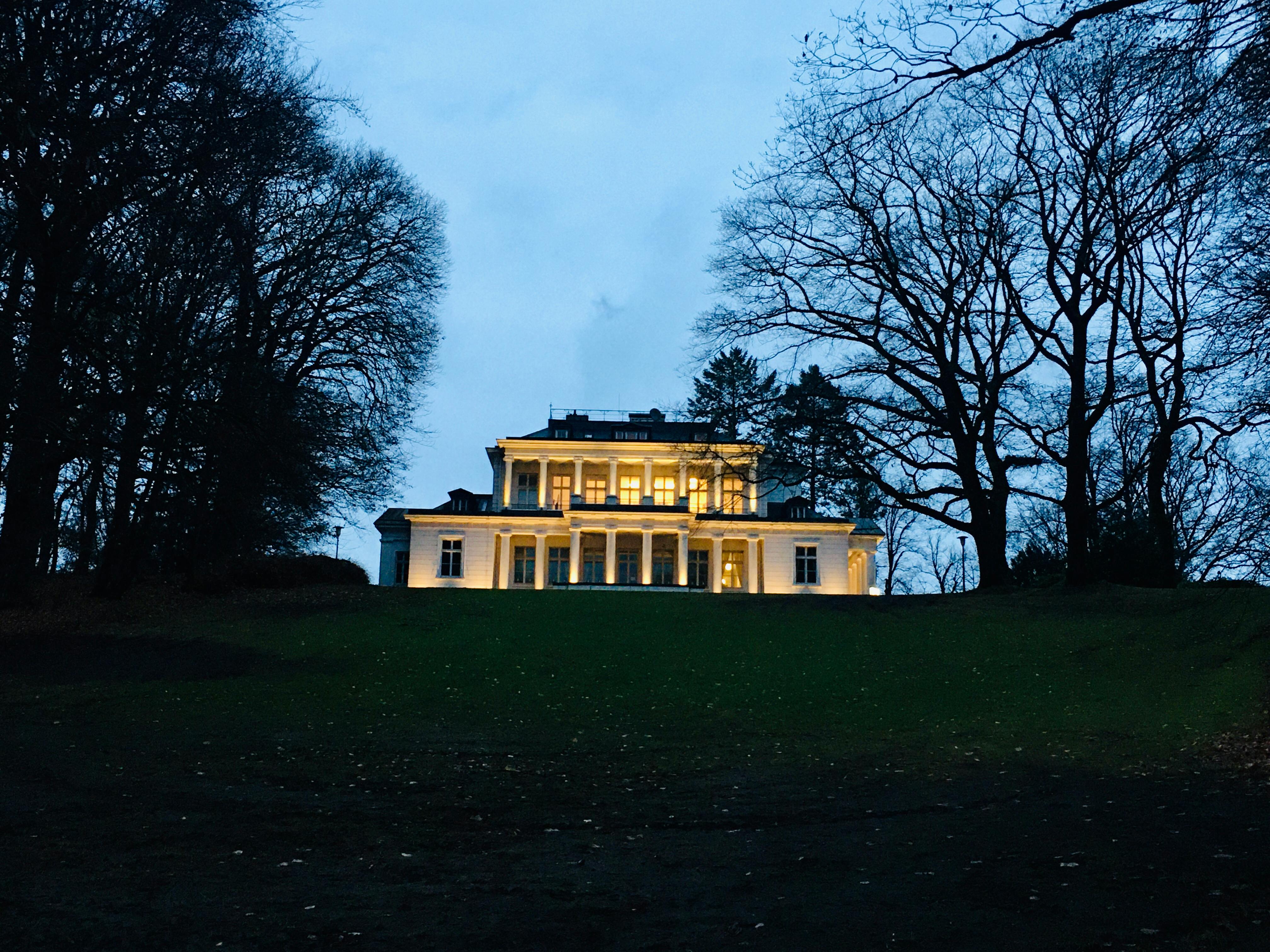 Eine angestrahlte alte Säulenvilla in einem Park mit Bäumen.