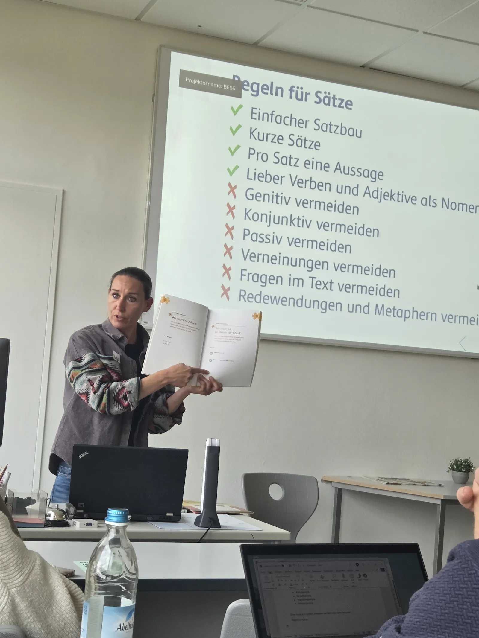 Nadja Quirein steht vor einem Whiteboard mit den Regeln der leichten Sprache und hält ein Buch in der Hand, in welchem die Regeln stehen.