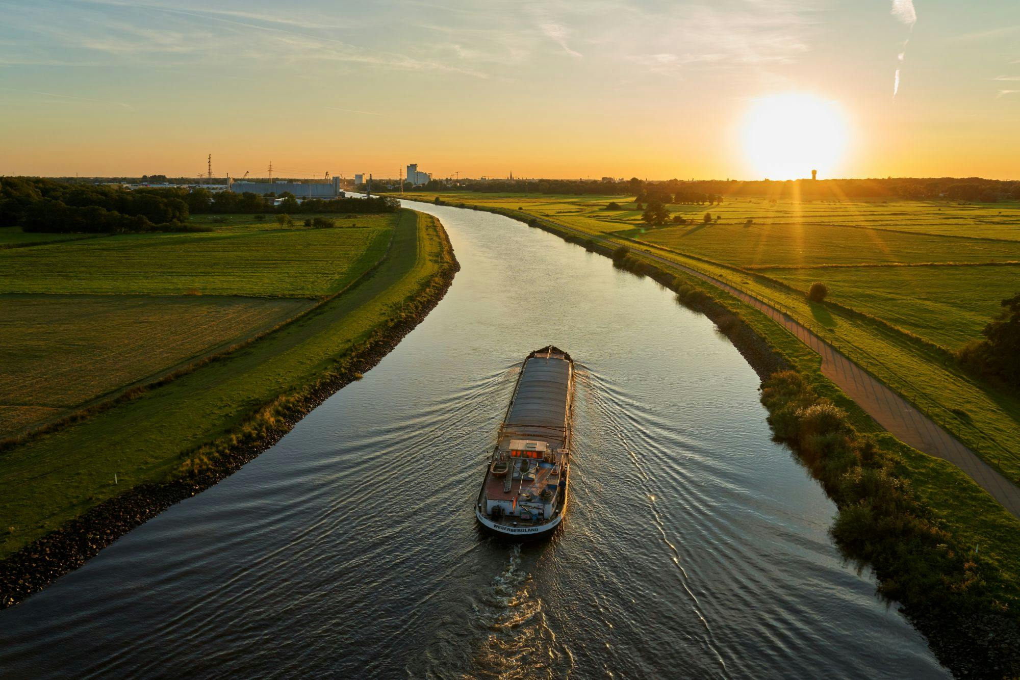 Der Fluss Hunte bei Oldenburg. Ein Kahn fährt im Sonnenuntergang darauf entlang. Im Hintergrund erkennt man die Skyline der Stadt.