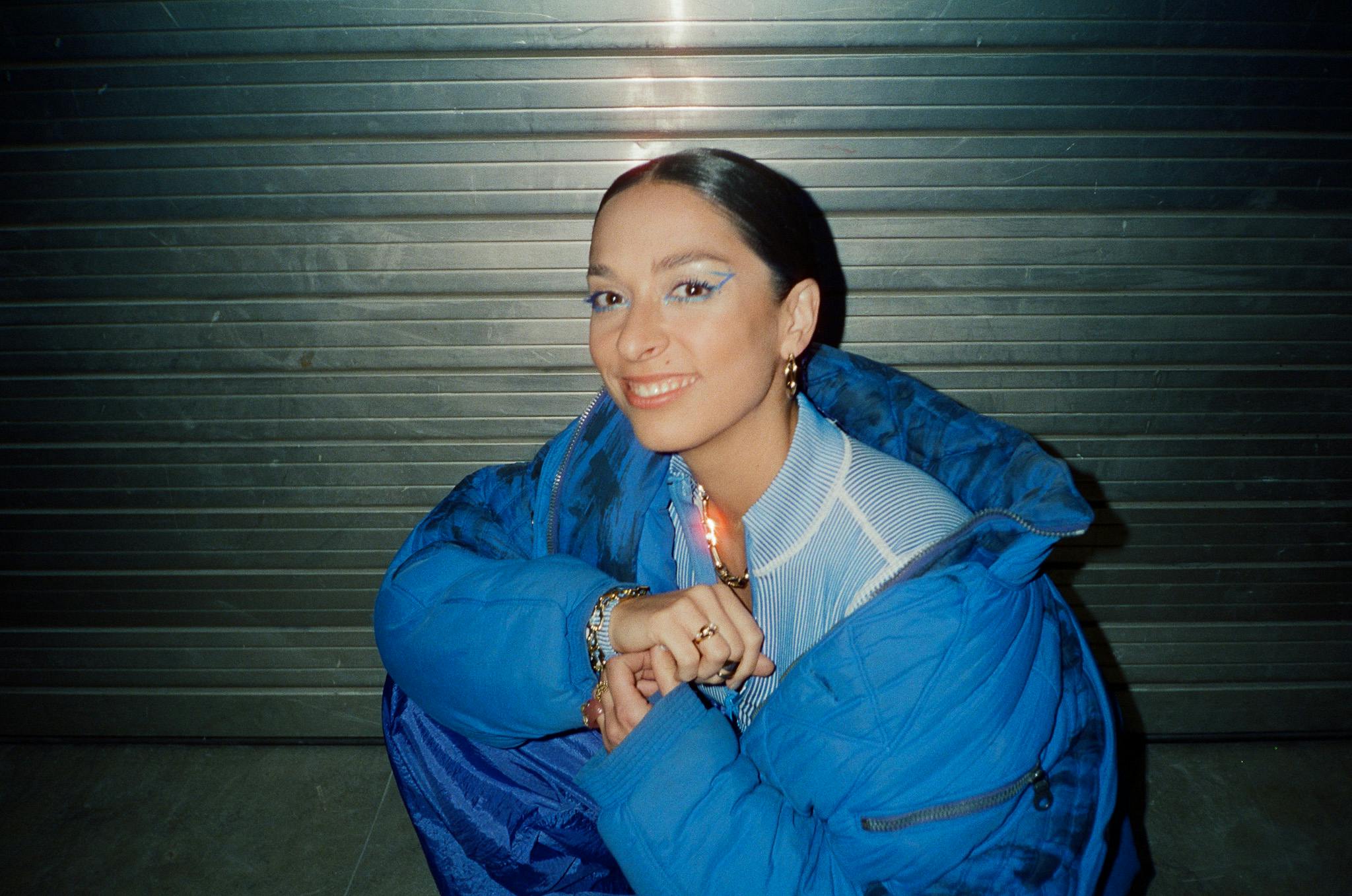 Tamara Güçlü aka Tam hockt im Blitzlicht mit himmelblauer Jacke vor einem Garagentor und lächelt.