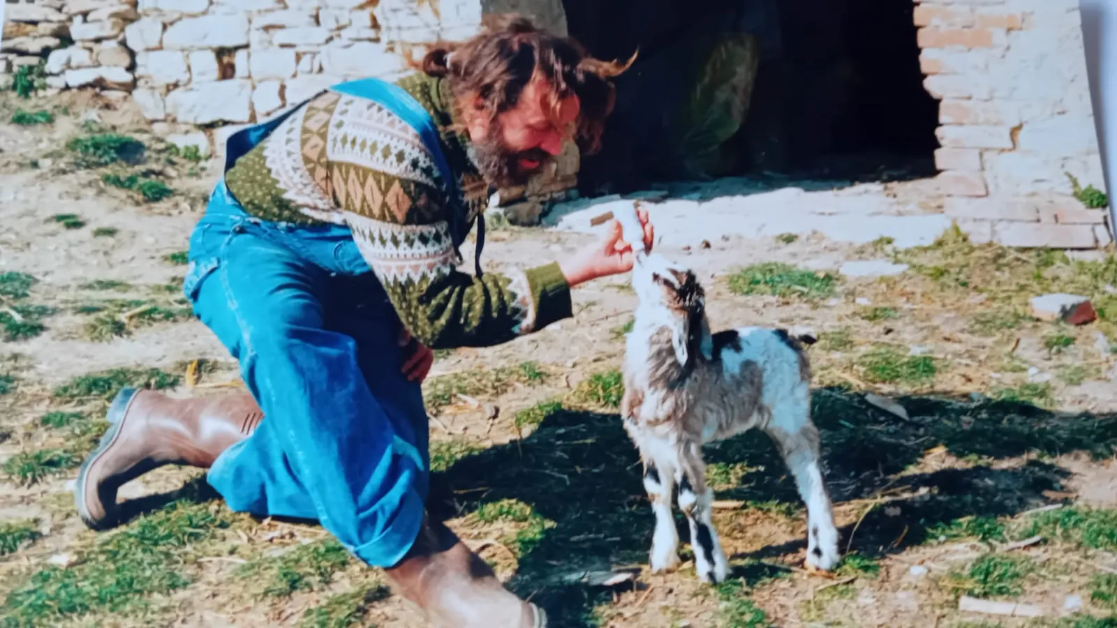 Uomo con salopet e stivali di gomma che da un biberon ad un agnellino, fuori da una stalla