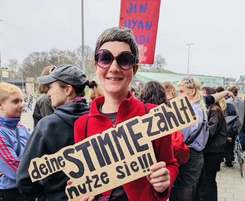Ein Foto von Ilona inmitten von rot-lila Demonstrantinnen, mit einem Schild in der Hand "Deine Stimme zählt – nutze sie!"