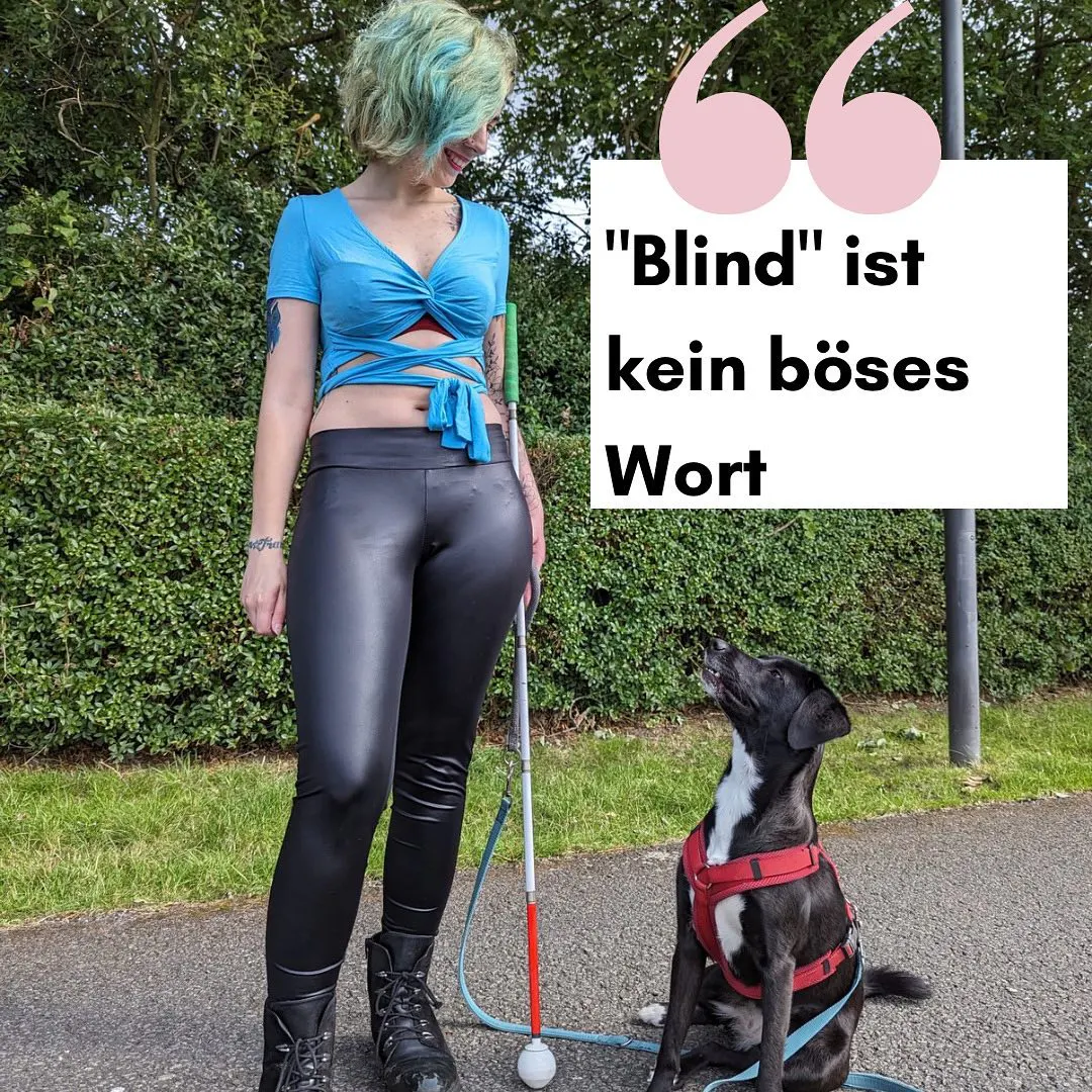 Nadine steht mit ihrer schwarz-weißen Hündin auf einem Weg. Im Hintergrund ist grün zu sehen. Nadine trägt schwarze Boots eine schwarze Leggins und ein blaues bauchfreies T-Shirt. Sie hat grüne Haare. Ihr Hündin sitzt neben ihr. Sie trägt ein rotes Geschirr. Sie schaut zu Nadine hoch. In einem weißen Kasten steht blind, ist kein böses Wort.