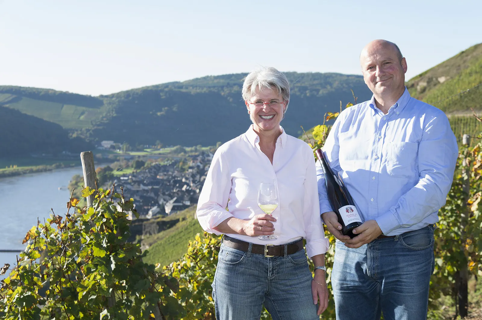 Barbara und Johannes Selbach vom Weingut Selbach-Oster stehen mit einer Weinflasche in ihrem Weinberg