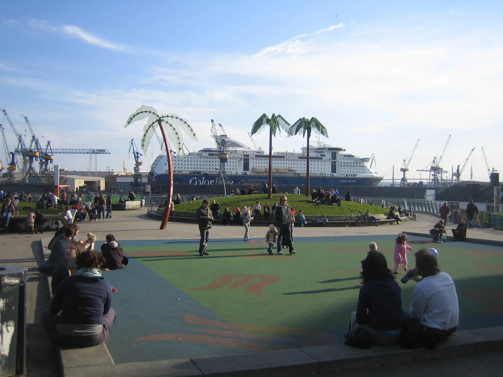 Park Fiction in St. Pauli. Menschen sitzen auf Kunststoffböden, Mauern und Bänken. Im Hintergrund fährt ein Kreuzfahrtschiff vorbei.