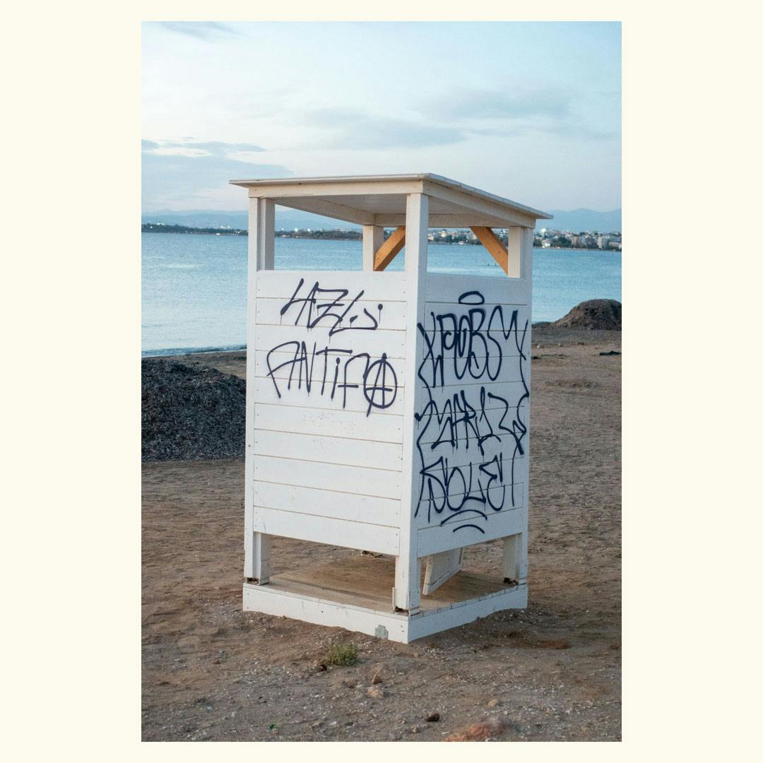Small white wooden structure with graffiti on two sides standing on a sandy beach with water and distant buildings in the background