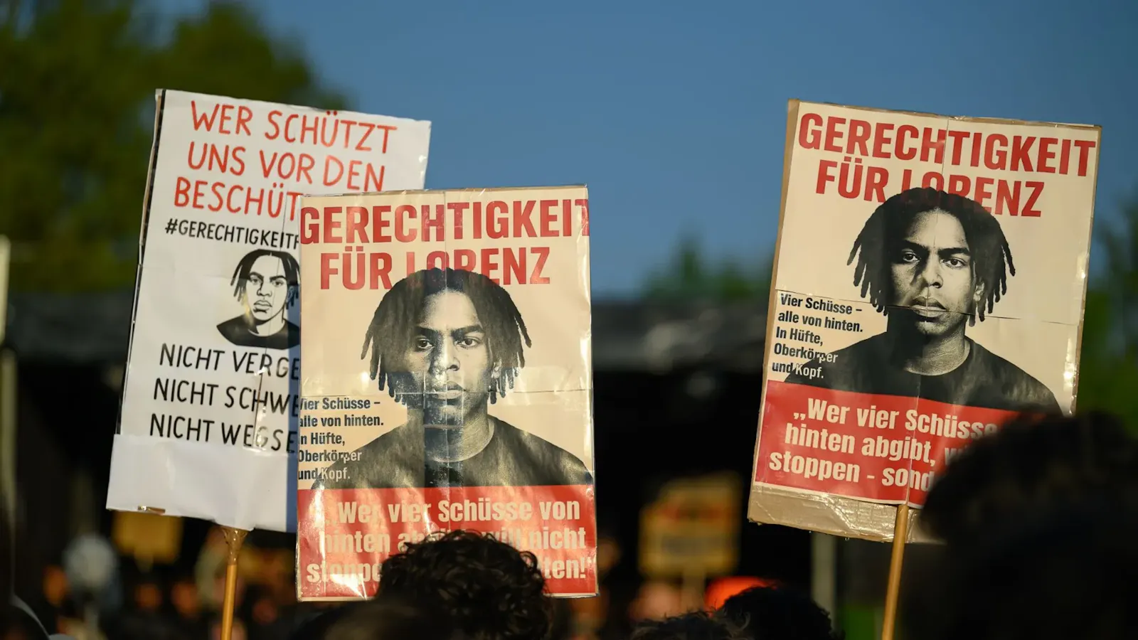 Bild von der Gedenkdemo für den erschossenen Lorenz. Es werden drei Plakate hochgehalten, die sein Konterfei zeigen, Text der vorderen beiden: "Gerechtigkeit für Lorenz - wer vier Schüsse von hinten abgibt, will nicht stoppen - sondern töten!" Text des hinteren Schildes: "Wer schützt uns vor den Beschützern? #gerechtigkeitfürlorenz nicht vergessen nicht schweigen nicht wegschauen"