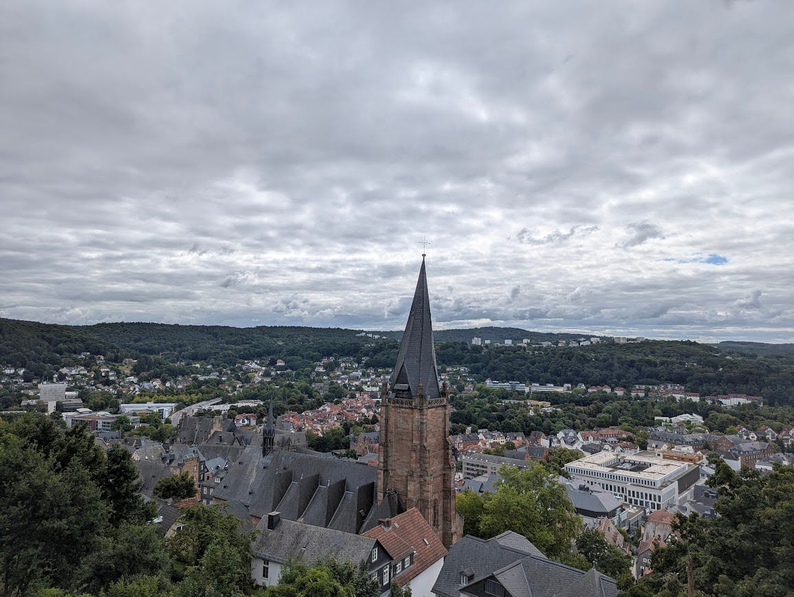 Blick von oben über Marburg und eine angrenzende Hügelkette; im Vordergrund eine auffällige Kirche mit leicht schiefem Kirchturm; stark bewölkter Himmel