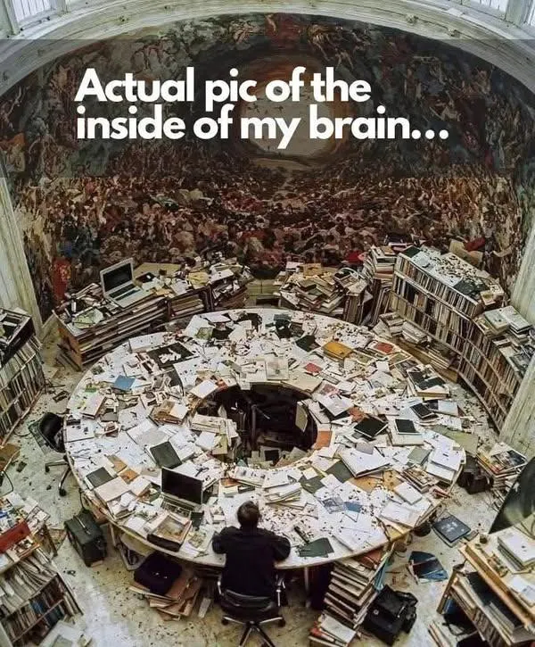 Ein Mensch sitzt an einem gut vier Meter durchmessenden runden Tisch voller Bücher, Zeitungen, Zettel und Monitore, umgeben von weiteren Stapeln Bücher, Zeitungen und Zettel.