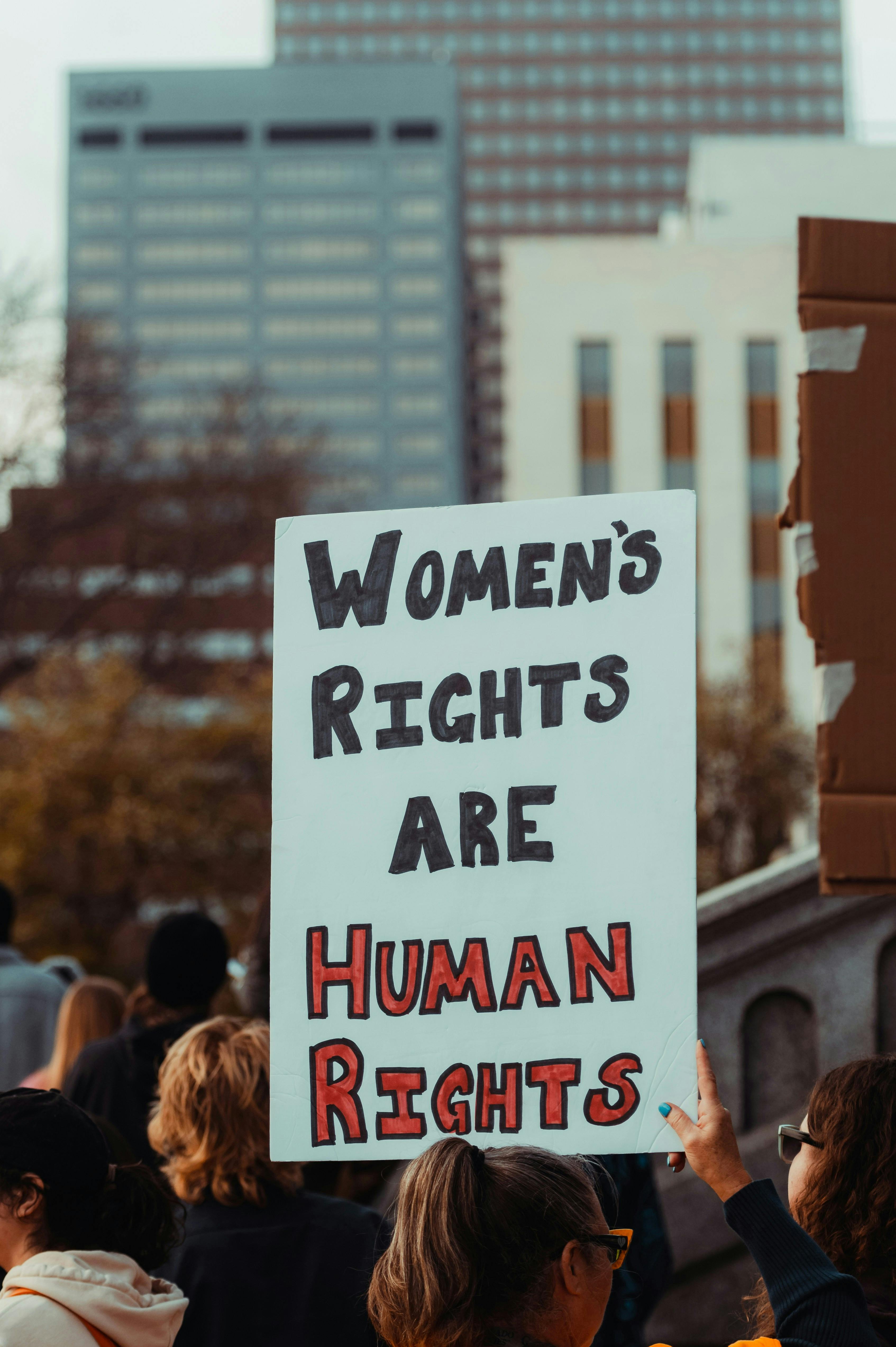 Ein Protestmarsch in einer Großstadt, eine Frau ist von hinten zu sehen. Sie trägt ein Schild, auf dem steht: „Women's Rights are Human Rights“.