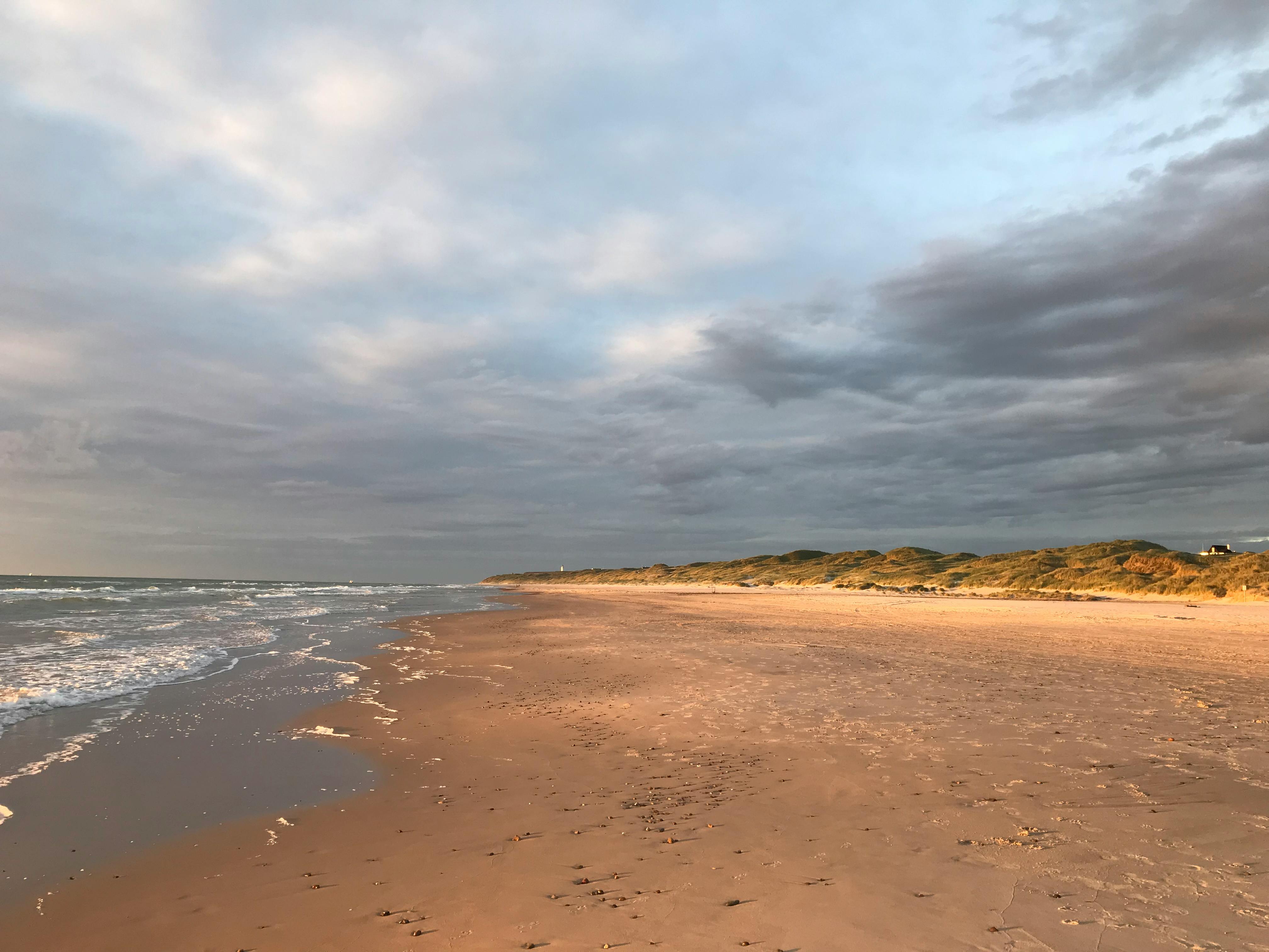 am meer. links die brandung, rechts grasbewachsene dünen, in denen ein haus zu erkennen ist. dazwischen ein breiter sandstrand. warmes abendlicht, ein weiter himmel voller wolkenformen und verschiedener blautöne. / at the seaside. surf to the left, grassy dunes to the right, in which you can make out a house. in between a wide sandy beach. warm evening light, vast sky full of different kinds of clouds and tints of blue.