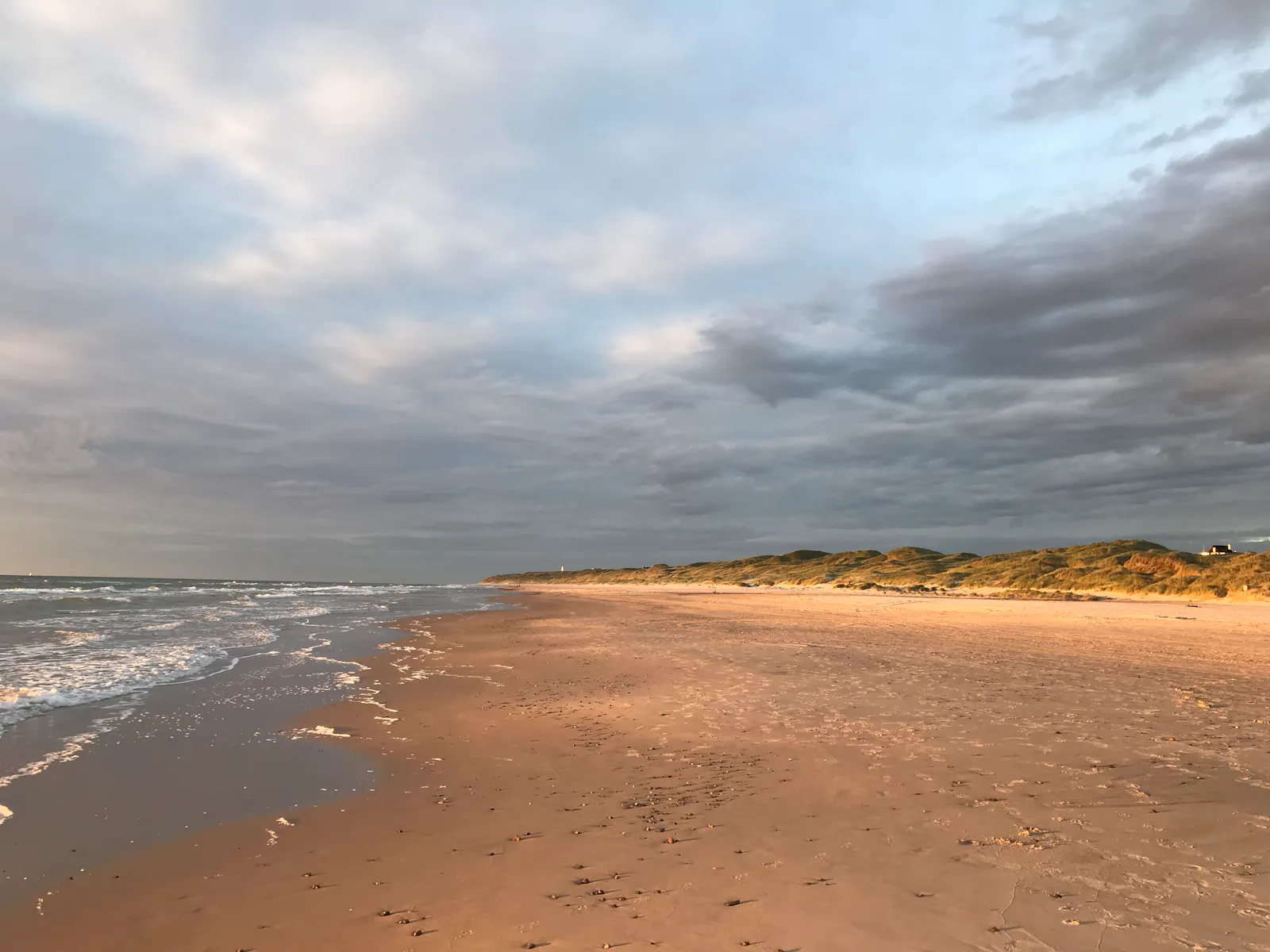 am meer. links die brandung, rechts grasbewachsene dünen, in denen ein haus zu erkennen ist. dazwischen ein breiter sandstrand. warmes abendlicht, ein weiter himmel voller wolkenformen und verschiedener blautöne. / at the seaside. surf to the left, grassy dunes to the right, in which you can make out a house. in between a wide sandy beach. warm evening light, vast sky full of different kinds of clouds and tints of blue.