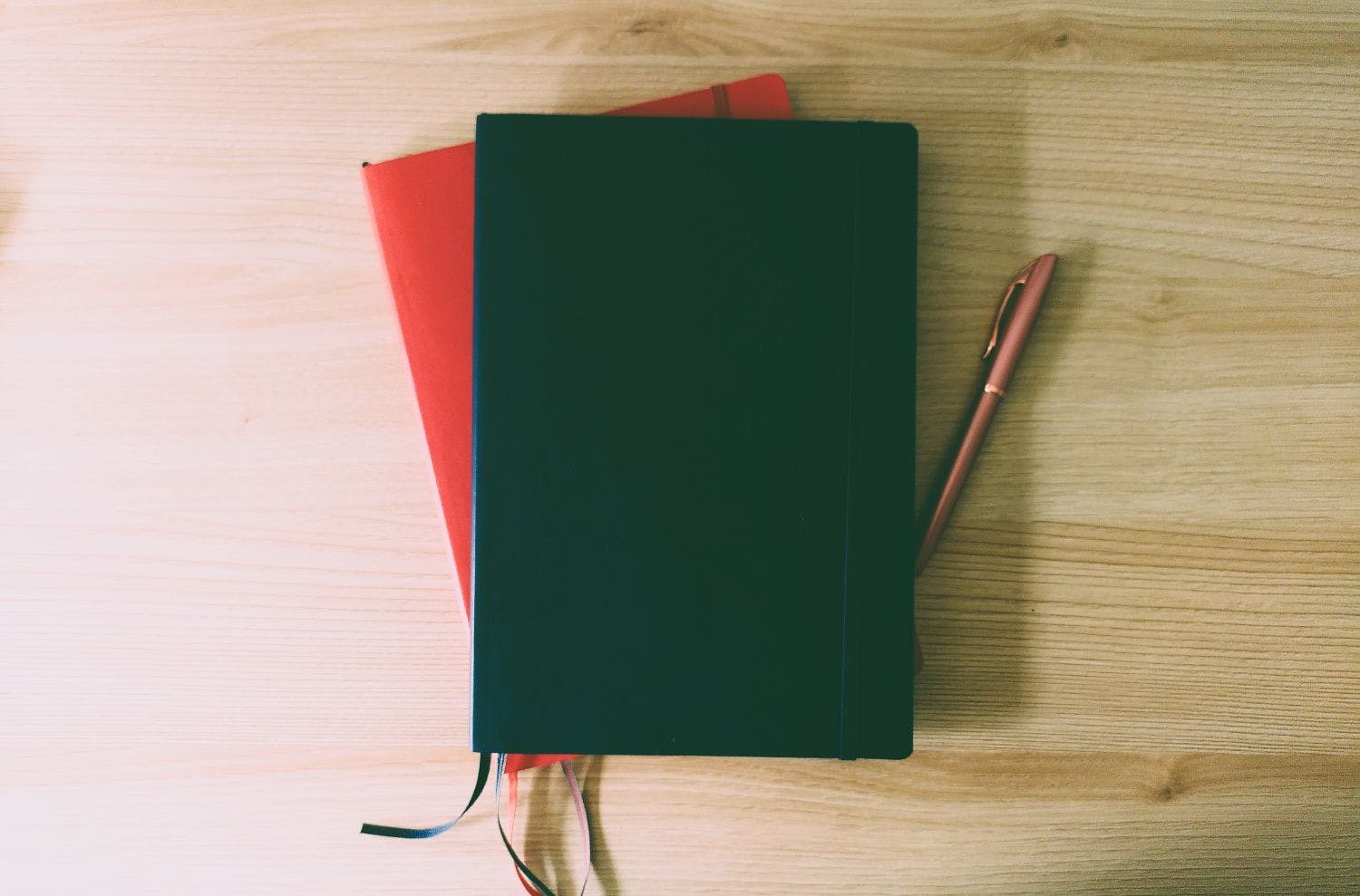 Zwei Notizbücher liegen auf einem Schreibtisch überinander, oben blau, unten rot. Rechts von den Notizbüchern liegt ein Füller.