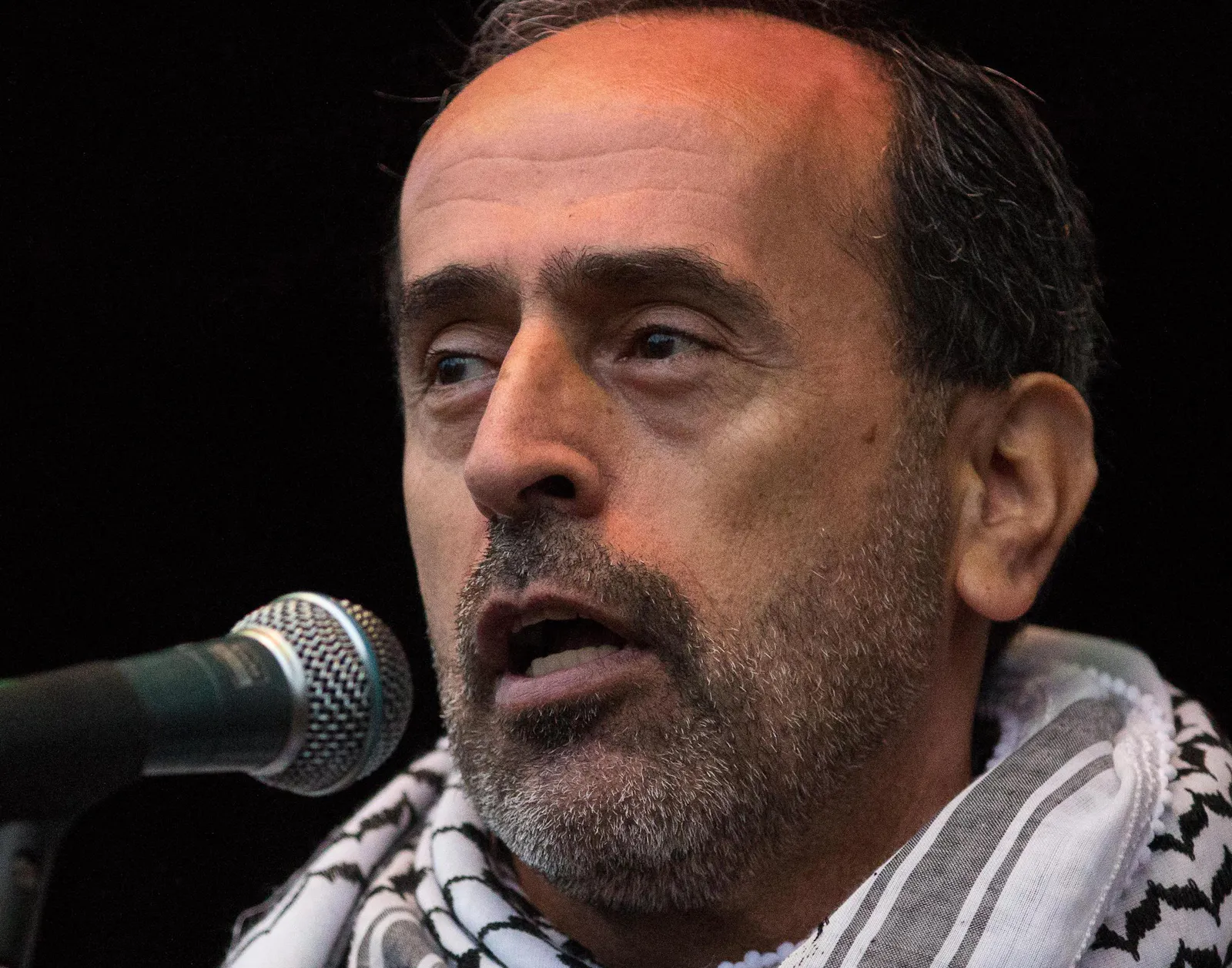 Zaher Birawi mit einer Kufiya auf den Schultern vor einem Mikrofon.