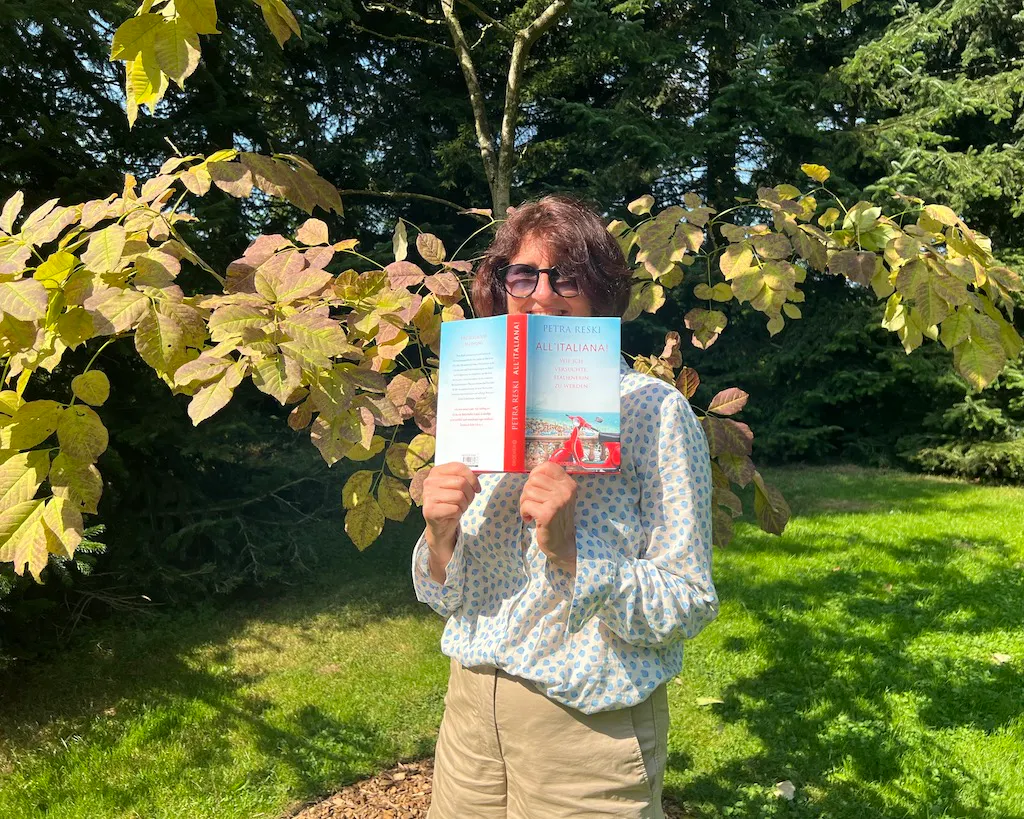 Ulrike Schmid steht in einem Park vor einem Laubbaum. Zu sehen ist ihr Oberkörper. In den Händen hält sie vor ihrem Gesicht das neueste Buch von Petra Reski, All'italiana. Wie ich versuchte, Italienerin zu werden. Ihr Gesicht ist halb vom aufgeklappten Buch zu sehen.