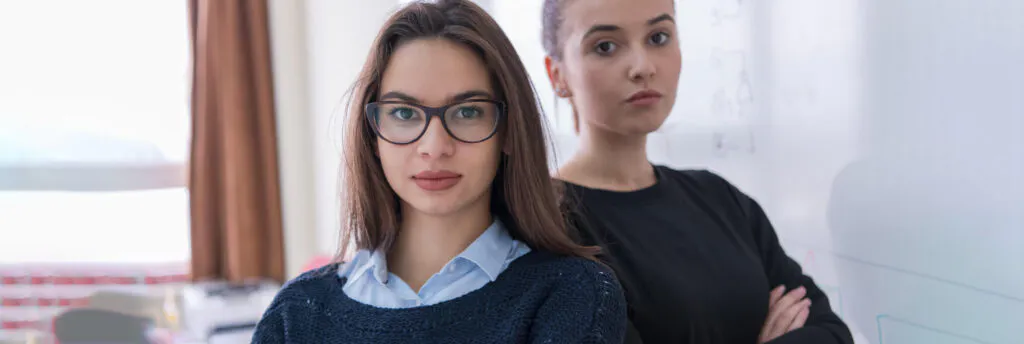 Zwei junge Frauen, die vor einer Tafel stehen und direkt in die Kamera schauen