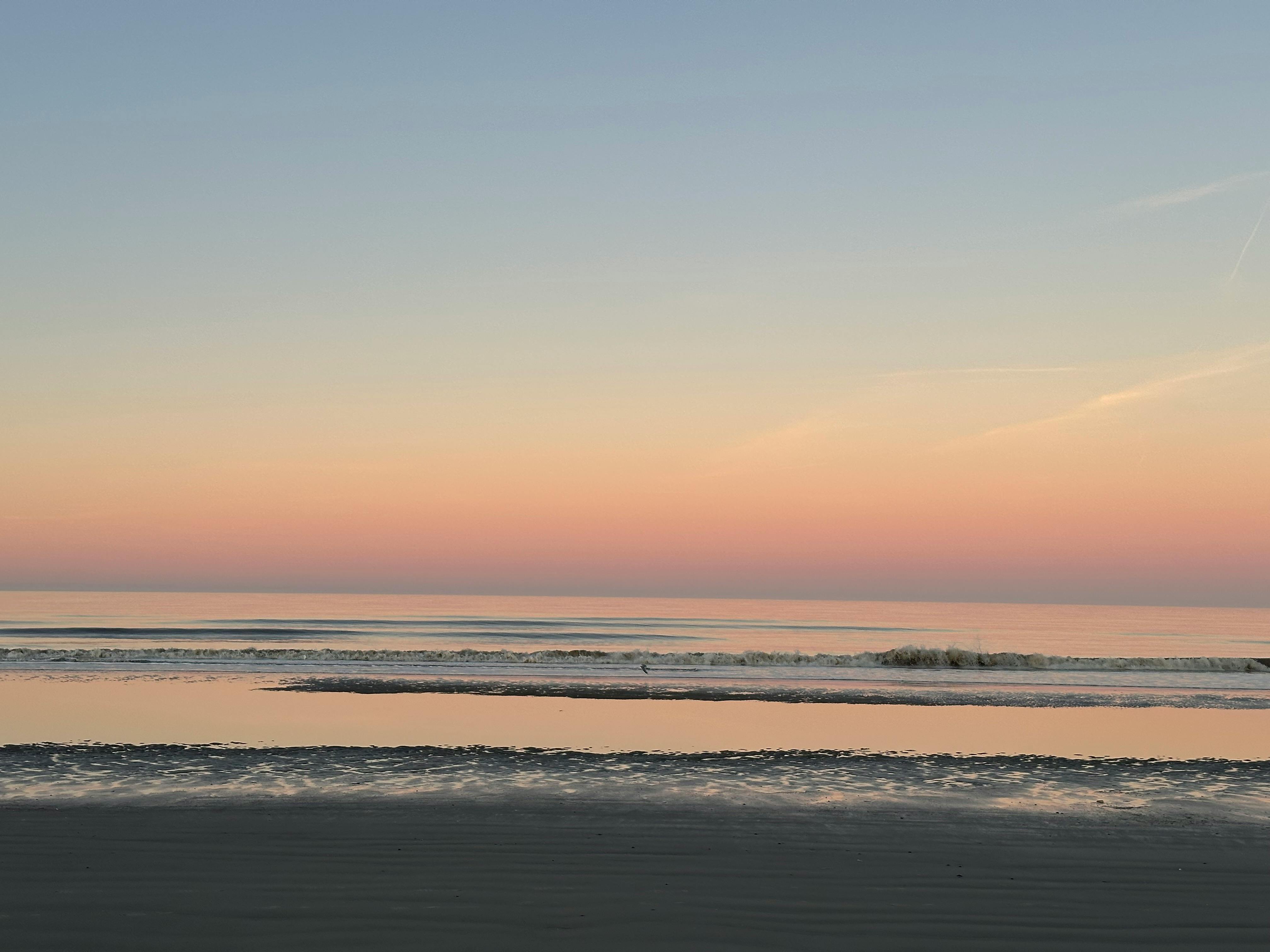 Meersaum bei Sonnenaufgang: Zartrosé- und hellblau-farbener Himmel, der sich im Meer spiegelt.