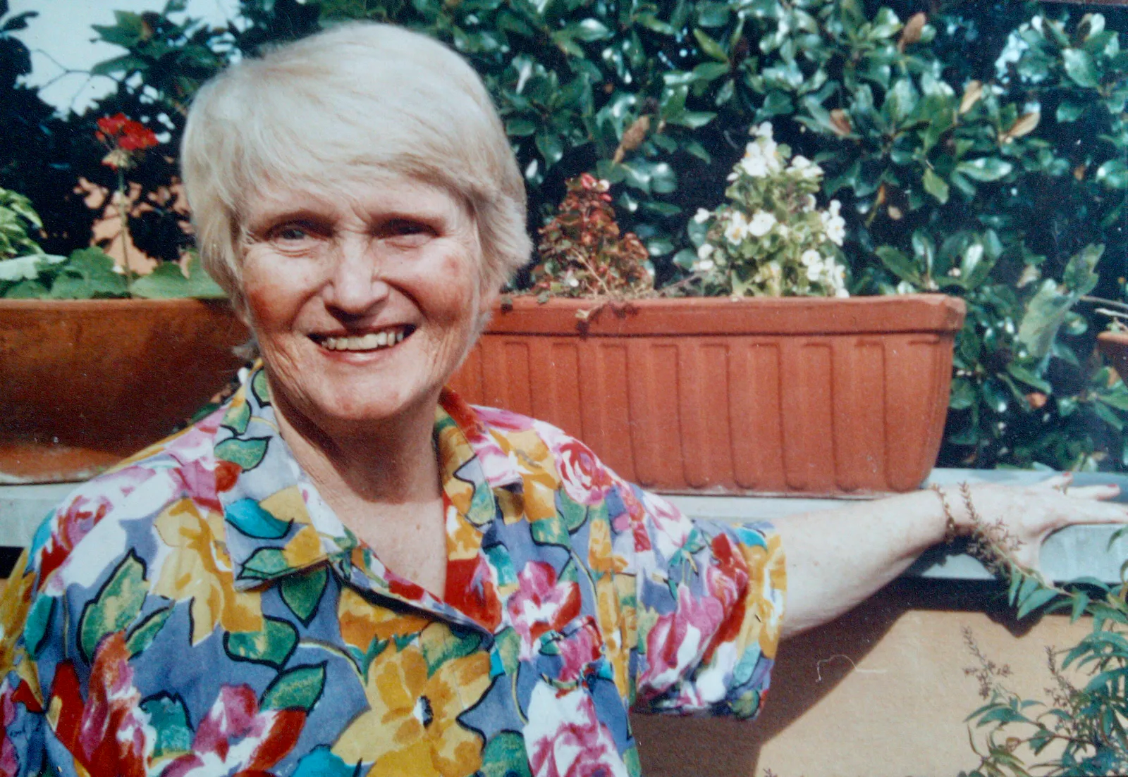 A woman with shortish blond hair and wearing a brightly coloured floral print shirt looks out at us smiling. Her left hand holds the wall of a terrace. Behind her are terracotta long vases with begonia, and way behind is a magnolia tree.