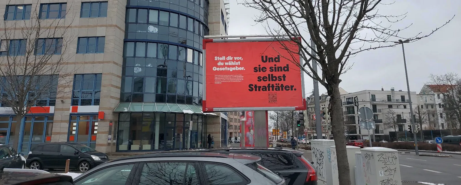 Das Bild zeigt eine Straßenansicht mit einem Werbeplakat im Vordergrund. Das Plakat hat einen roten Hintergrund und zeigt in großen schwarzen Buchstaben den Text: 'Stell dir vor, du wählst Gesetzgeber. Und sie sind selbst Straftäter.' Rechts unten auf dem Plakat befindet sich ein QR-Code. Im Hintergrund sieht man ein modernes Gebäude mit großen Fenstern und parkende Autos. Der Himmel ist bewölkt, und auf den Straßenbäumen sind keine Blätter mehr zu sehen, was auf den Winter hinweist.