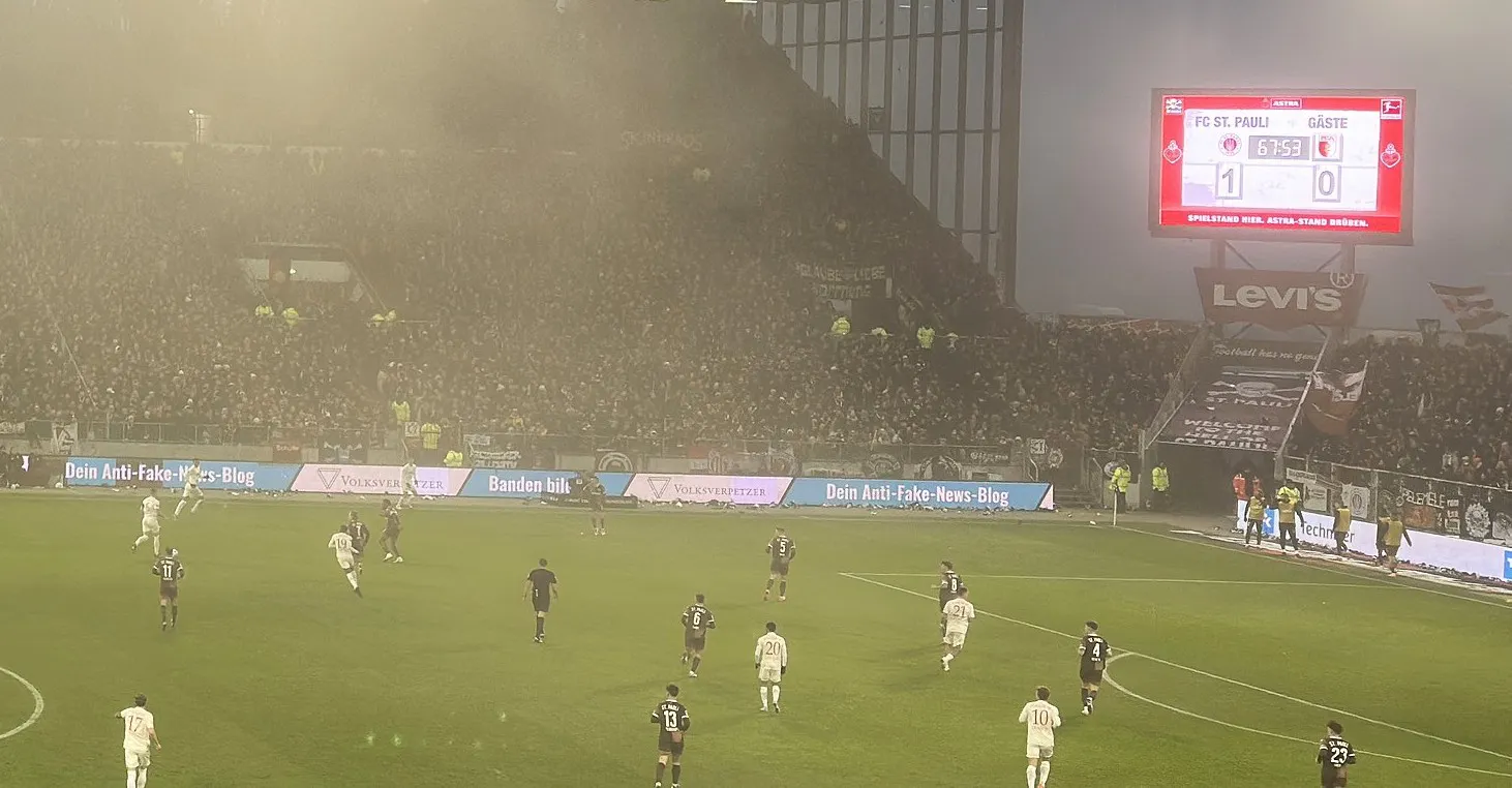 Das Bild zeigt ein Fußballstadion während eines Spiels. Auf dem Spielfeld sind Spieler beider Mannschaften zu sehen. Im Hintergrund ist eine große, dicht besetzte Fankurve, die durch Nebel oder Dunst etwas verschwommen wirkt. Der Bildschirm im Stadion zeigt den aktuellen Spielstand: FC St. Pauli führt mit 1:0 gegen die Gäste. Auf den Werbetafeln rund um das Spielfeld sind die Schriftzüge 'Volksverpetzer', 'Dein Anti-Fake-News-Blog' sowie die Nachricht 'Banden bilden!' zu sehen.