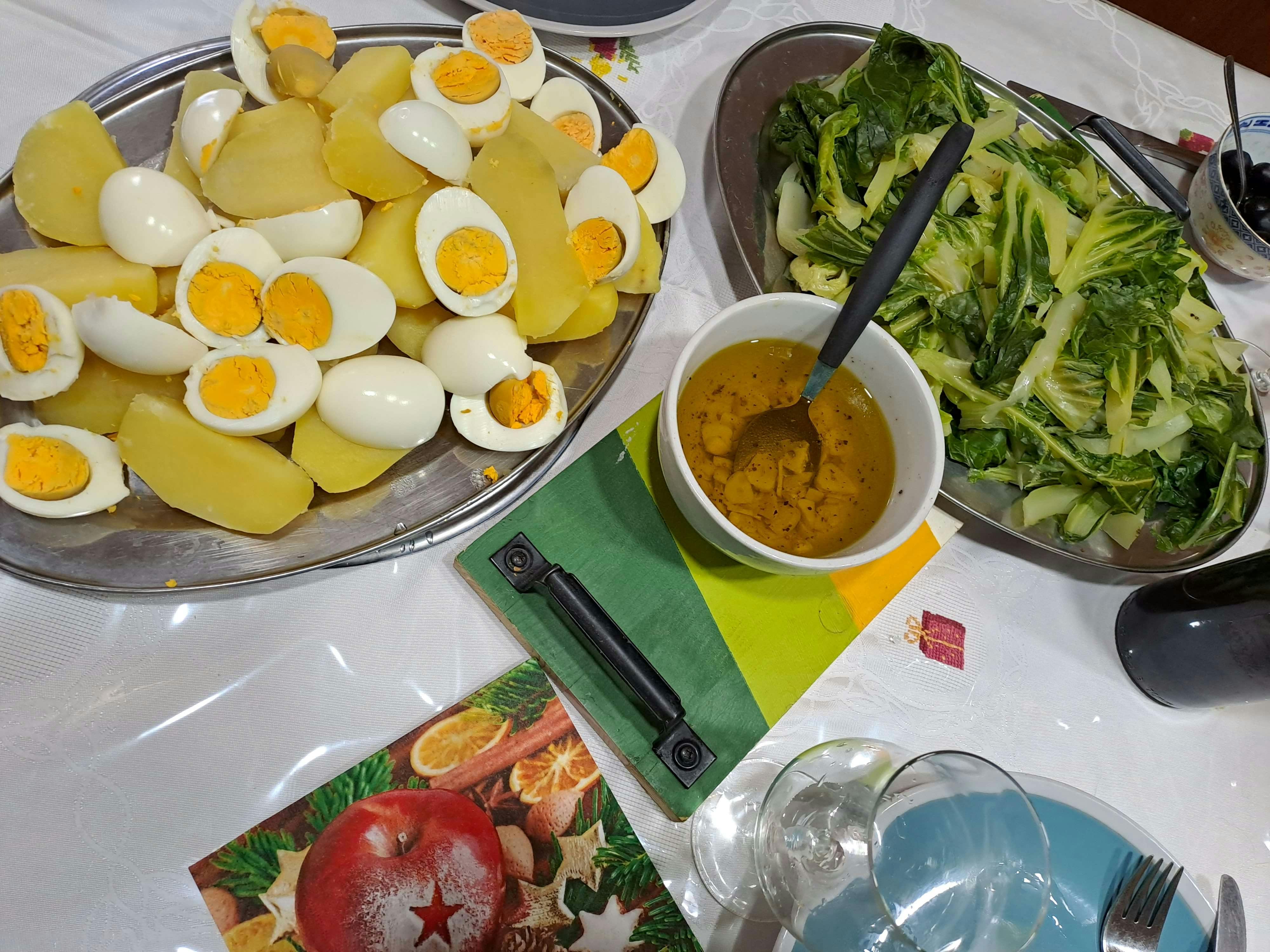 Festlich gedeckter Tisch mit einem portugiesischen Weihnachtsgericht: gekochte Kartoffeln und halbierte Eier auf einer Platte, daneben eine Schüssel mit grünem Kohl, eine kleine Schale mit Öl-Knoblauch-Dressing sowie Teller, Besteck und Gläser.
