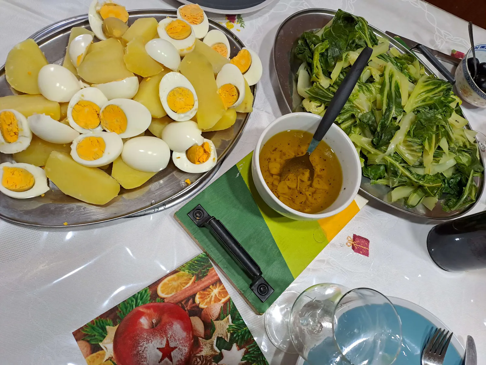 Festlich gedeckter Tisch mit einem portugiesischen Weihnachtsgericht: gekochte Kartoffeln und halbierte Eier auf einer Platte, daneben eine Schüssel mit grünem Kohl, eine kleine Schale mit Öl-Knoblauch-Dressing sowie Teller, Besteck und Gläser.