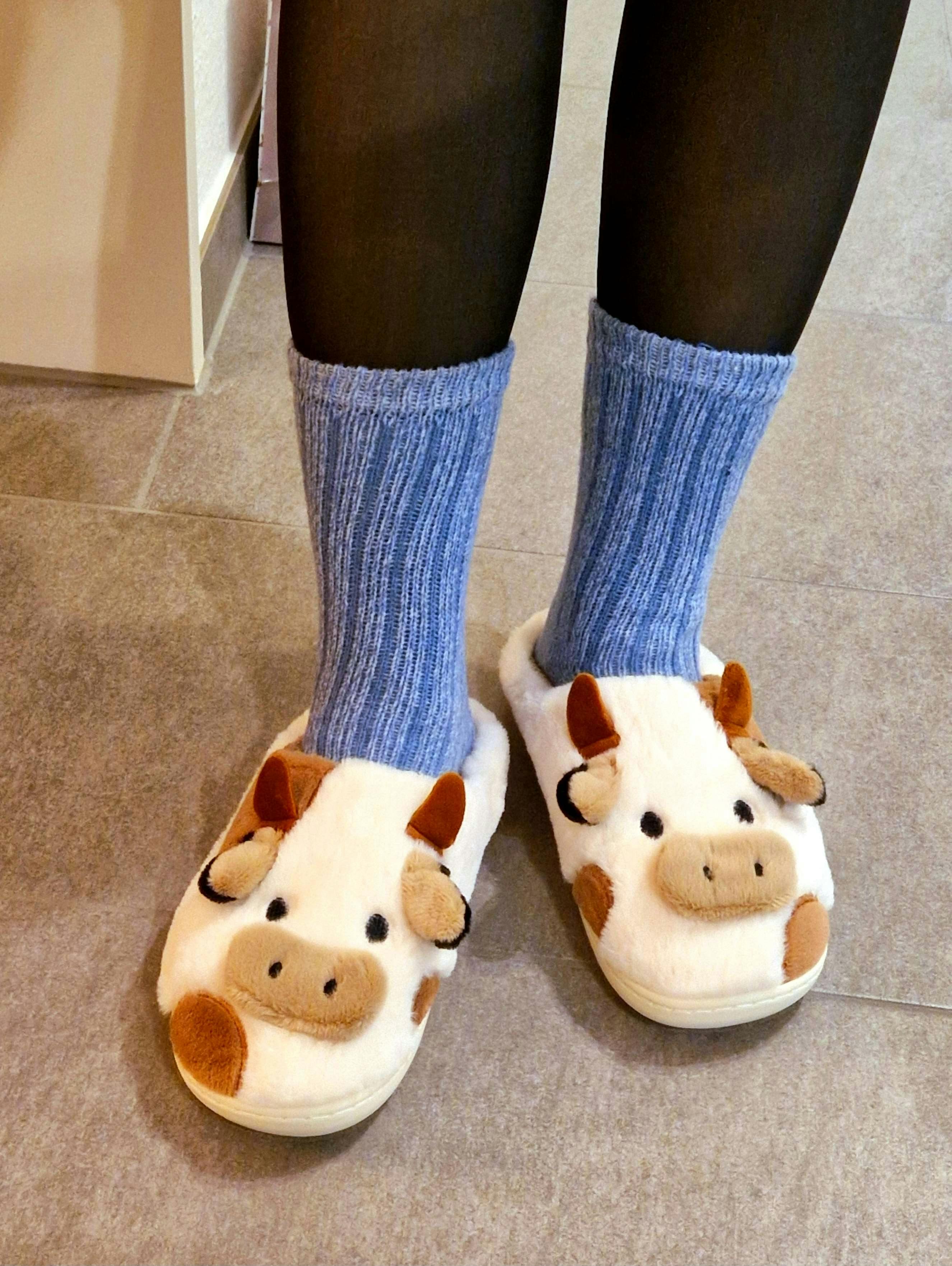 Skizzy steht auf Fliesenboden und trägt warme Socken sowie flauschige Hausschuhe in Kuhform mit kleinen Hörnern und aufgesticktem Gesicht.