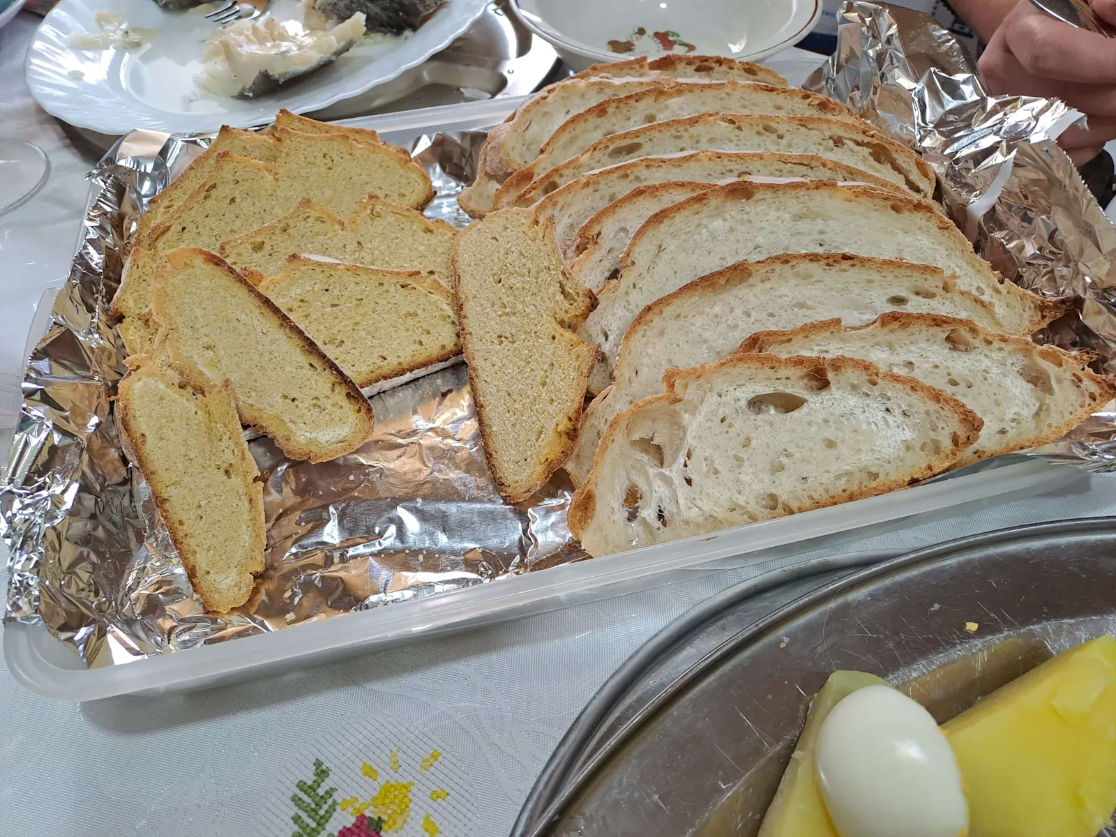 Aufgeschnittenes portugiesisches Brot und helles Gebäck in Scheiben, auf Alufolie in einer Kunststoffbox angerichtet.