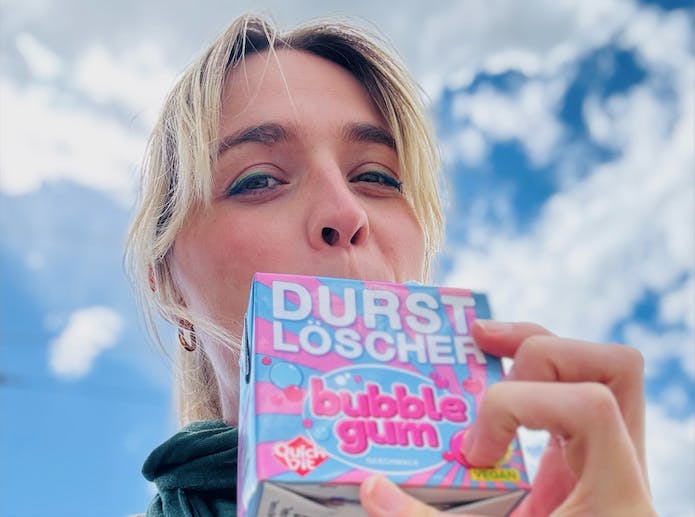 Auf diesem Selfie ist Nyke Slawik mit einem Bubble Gum Durstlöscher zu sehen der den Mund verdeckt. Im Hintergrund ist blauer Himmel und Wolken zu sehen.