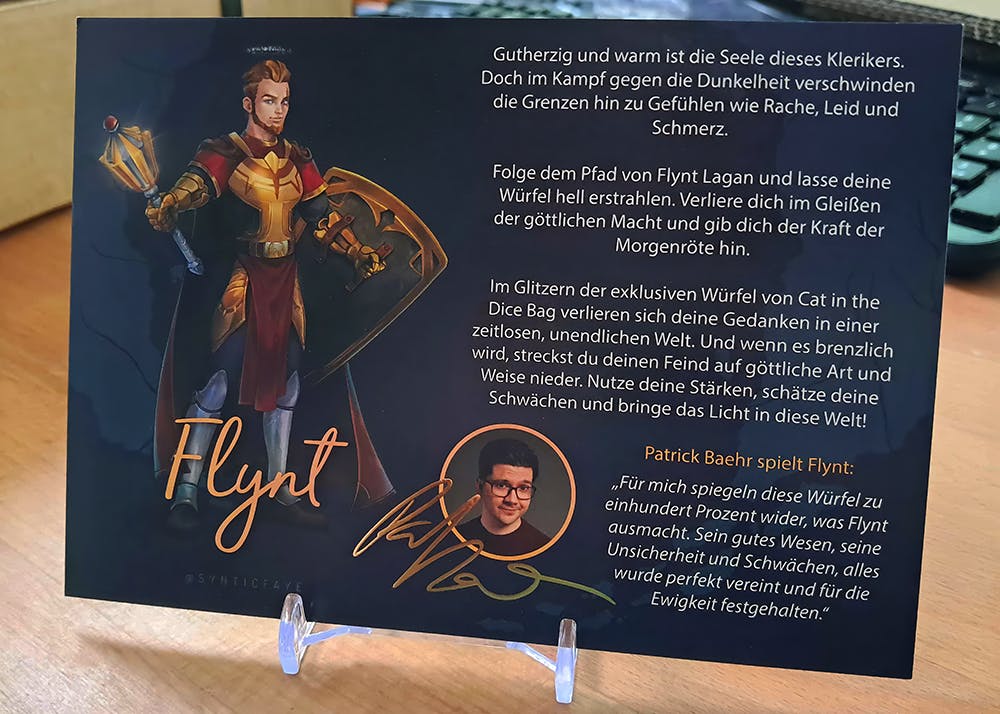 Eine Karte auf der Flynt abgebildet ist. Daneben ein Text über die Würfel und Patzes Gedanken zu den Würfeln. Außerdem ein kleines Foto von Patze und eine goldene Unterschrift.