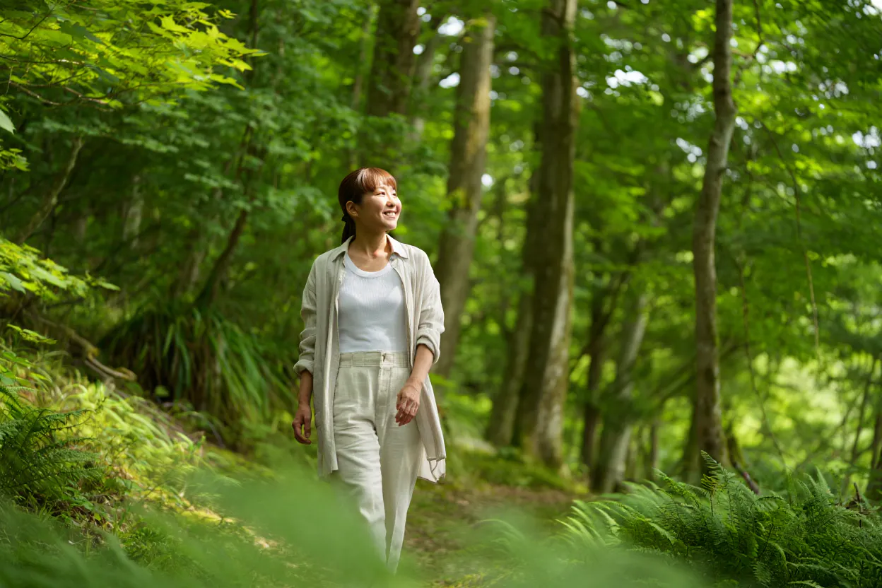 Eine Frau mittleren Alters läuft bei mit legerer heller Kleidung durch einen Wald. Sie hat einen zufriedenen Gesichtsausdruck