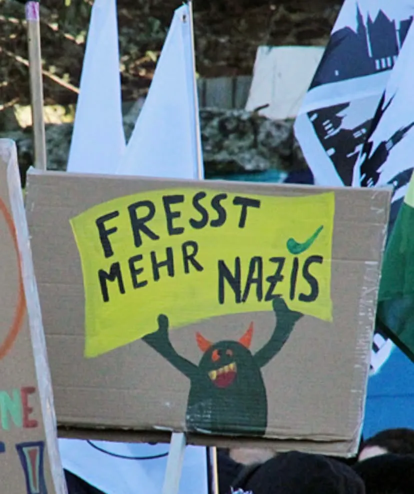 Nahaaufnahme eines Demoschilds aus Pappe, ein dunkelgrünes Monster mit orangenen Hörnern hält ein gelbfarbenes Schild hoch, auf dem "Fresst mehr Nazis" steht