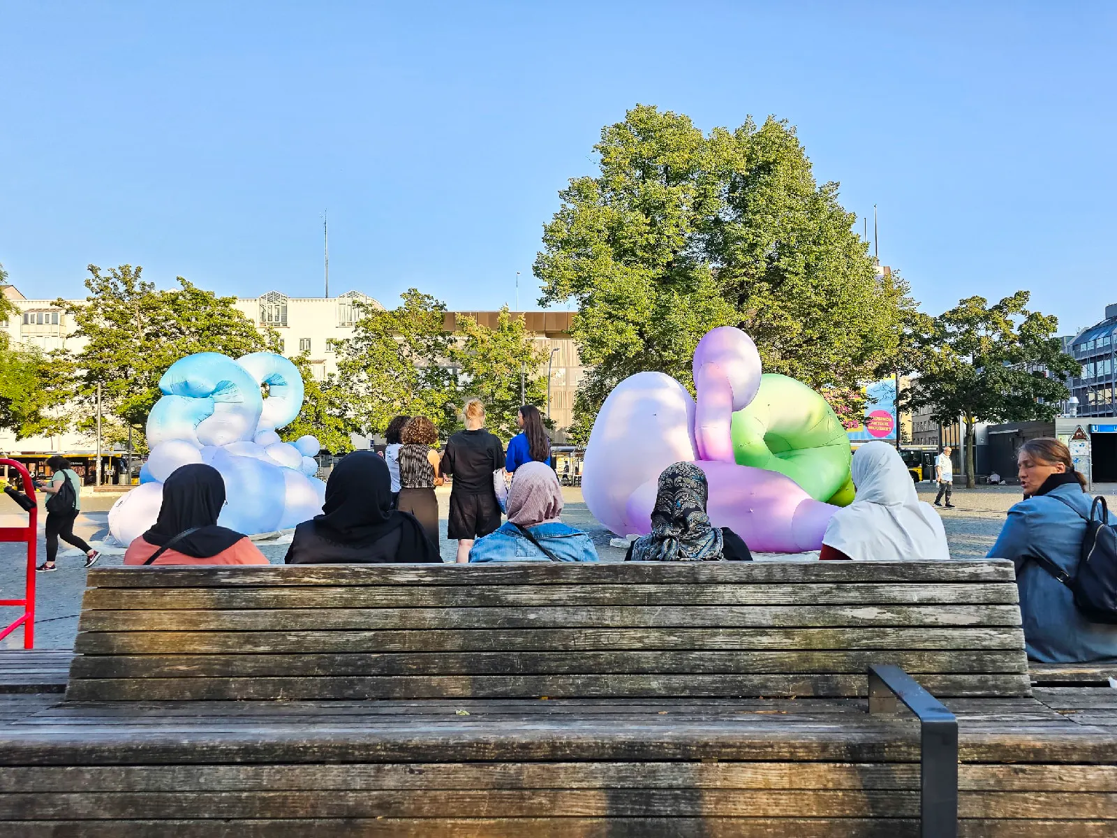 Fünf Frauen mit Kopftuch von hinten auf einer Bank, vor sich drei große amorphe, aufgeblasene Objekte in Blau, Lila, Grün. Zwischen Frauen und Objekten eine Gruppe von Frauen ohne Kopftuch, die uns ebenfalls den Rücken zuwenden. Baume, strahlend blauer Himmel.
