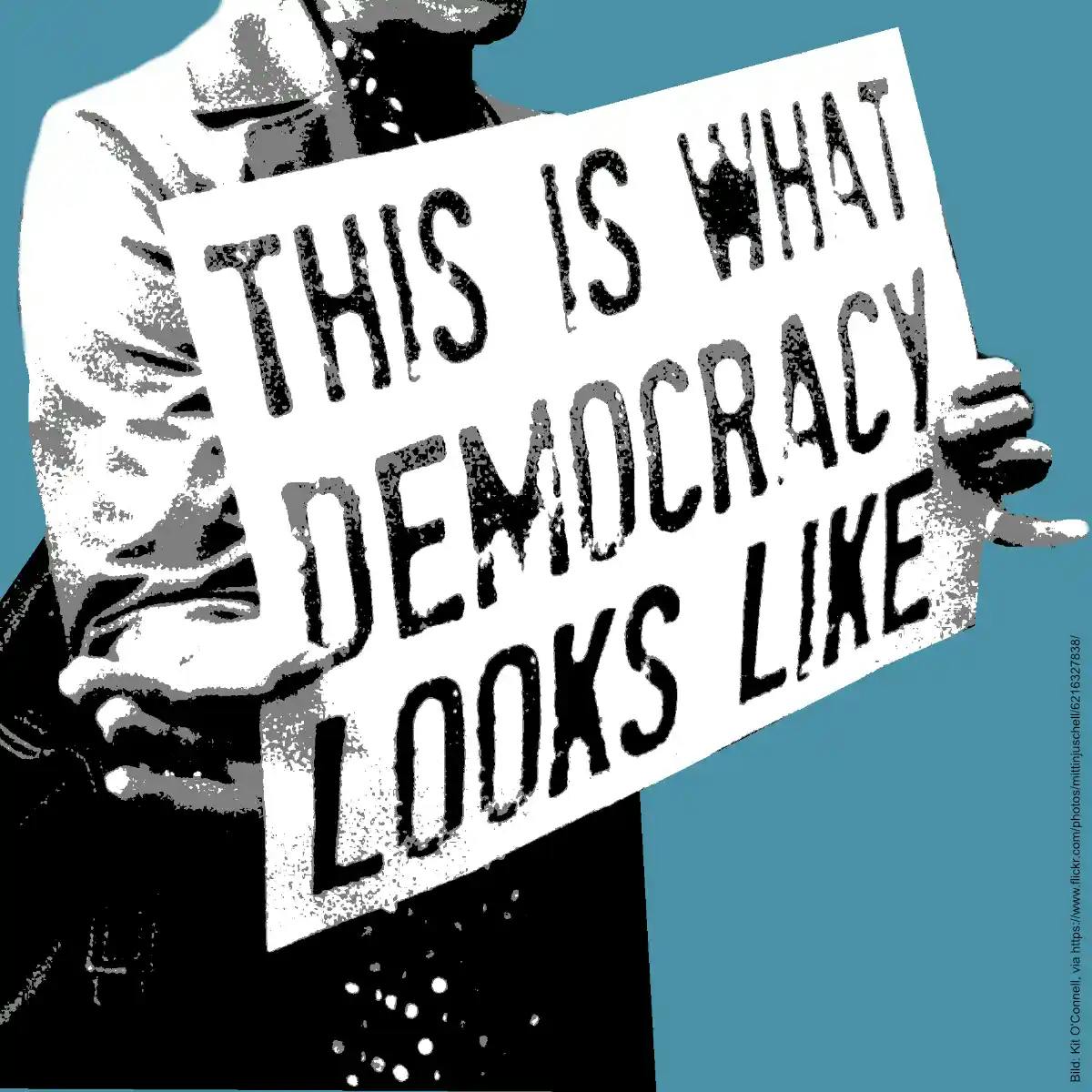 [Eine Frau hält ein Schild hoch, auf dem steht "This is what democracy looks like"]