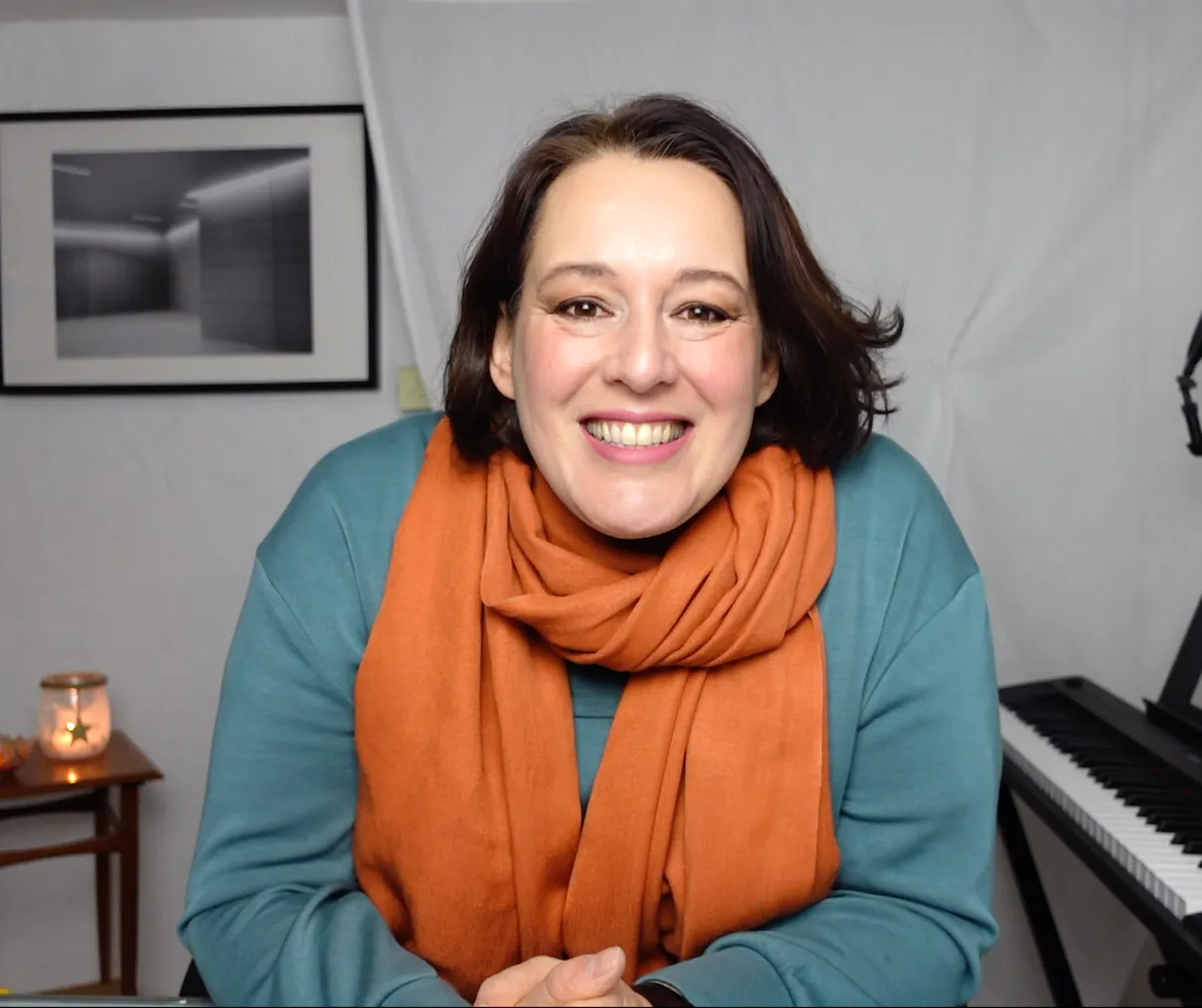Eine Frau mit dunklen, mittellangen Haaren lacht in die Kamera. Sie trägt einen orangenen Schal und einen Türkisen Pullover.