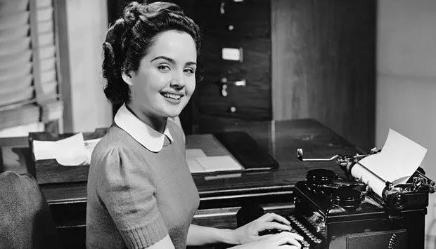 ein schwarz-weiß-Foto einer Sekretärin vor einer Schreibmaschine in einem klassischen 50er-Jahre-Stil lächelt in die Kamera.