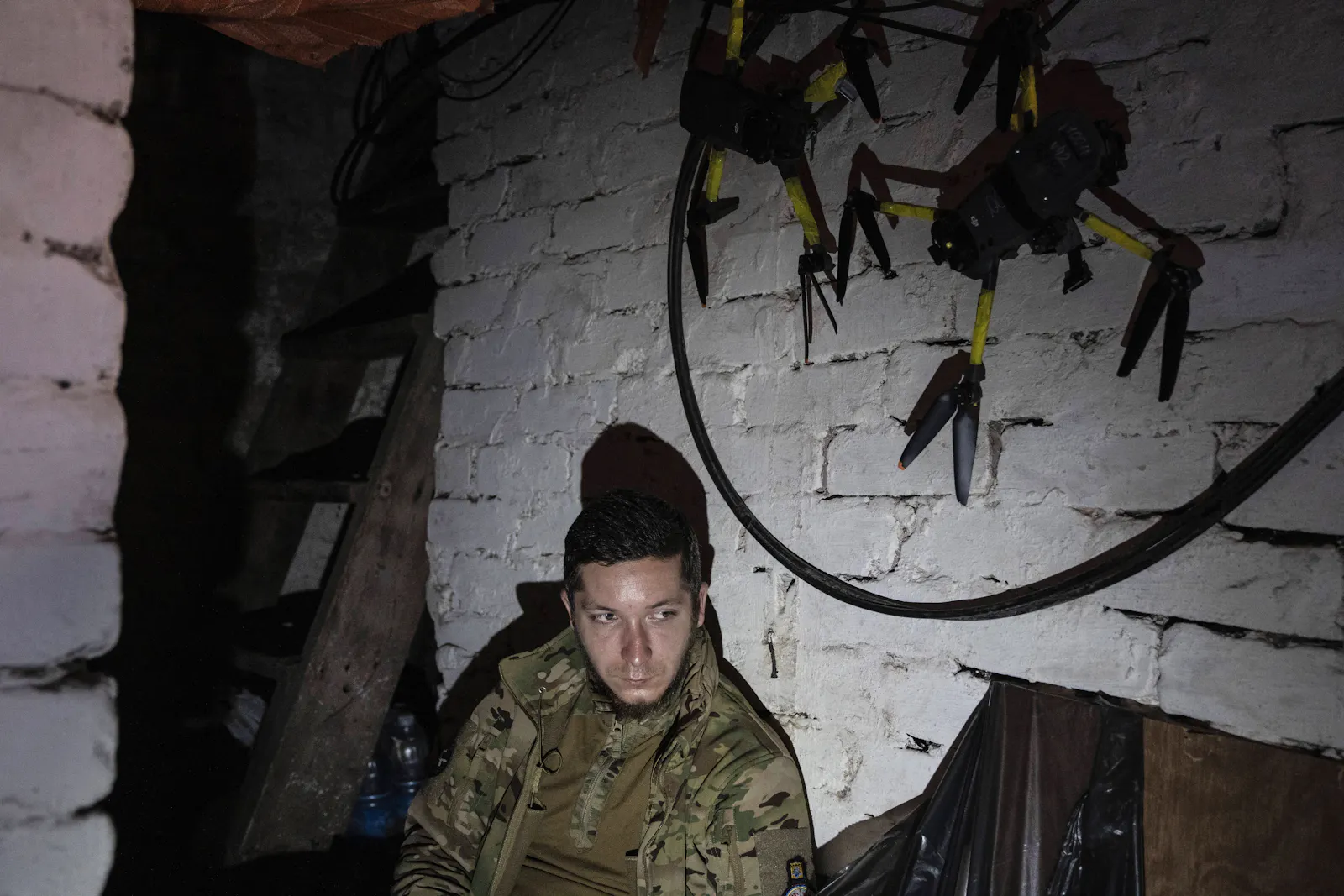 Ein junger urkainischer Soldat sitzt erschöpft in einem Keller, hinter ihm einige Drohnen.