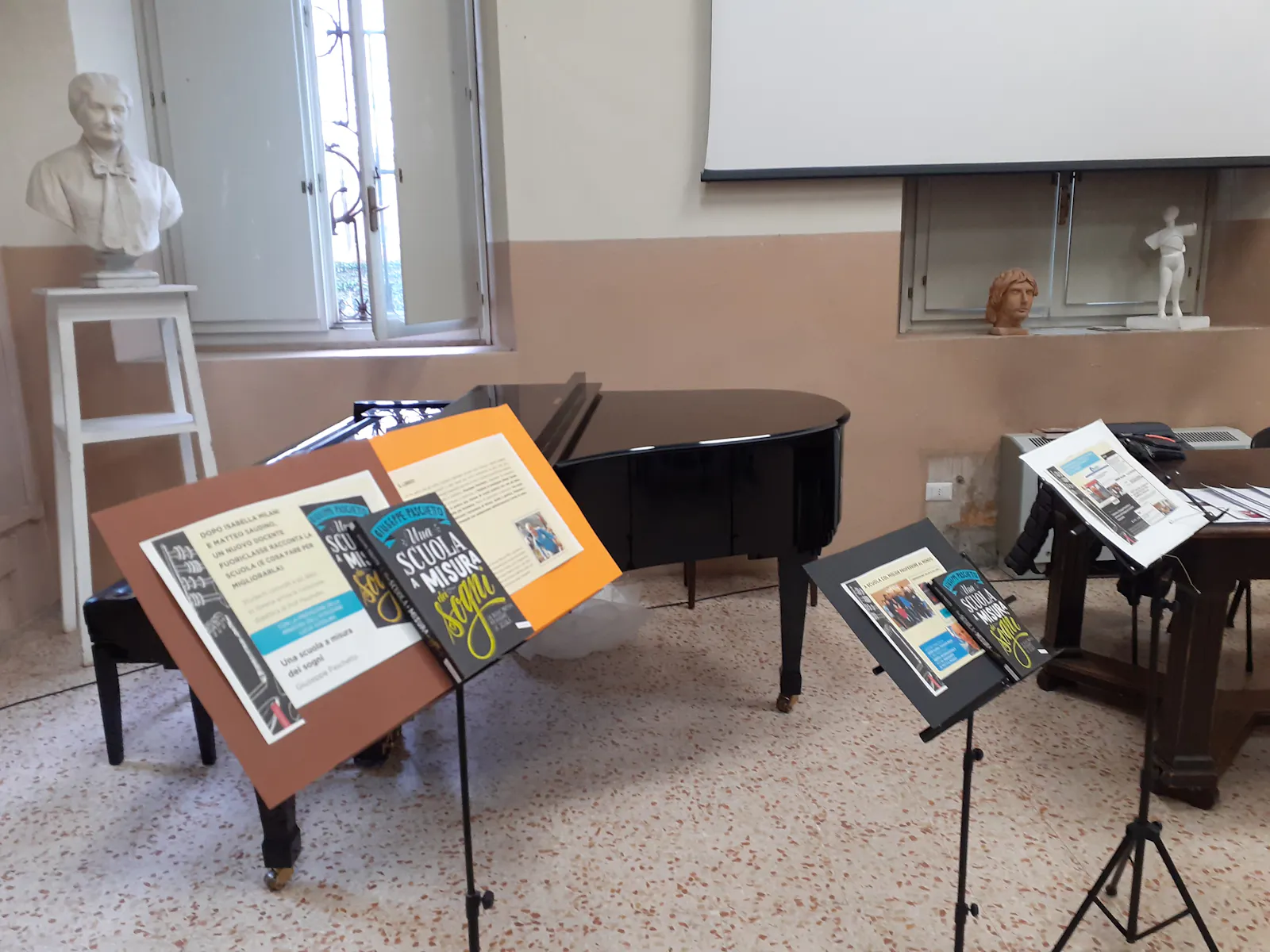 foto di un aula con pianoforte a mezzacoda e leggii su cui si vedonno più copie di "Una scuola a misura dei sogni2 di Giuseppe Paschetto