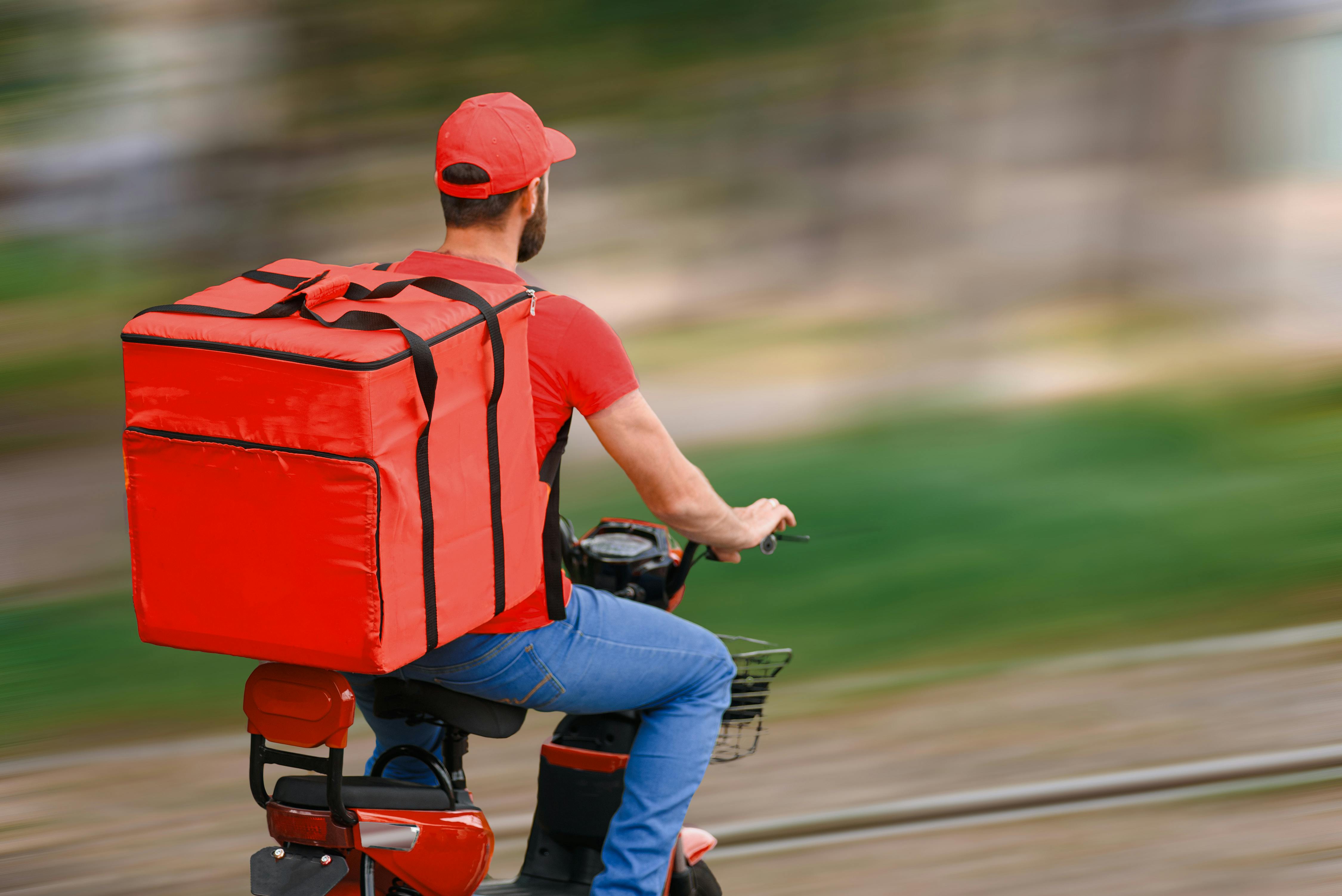 Das Bild zeigt einen Liefer-Fahrer auf einem E-Moped mit einem großen roten Thermo-Rucksack auf dem Rücken.