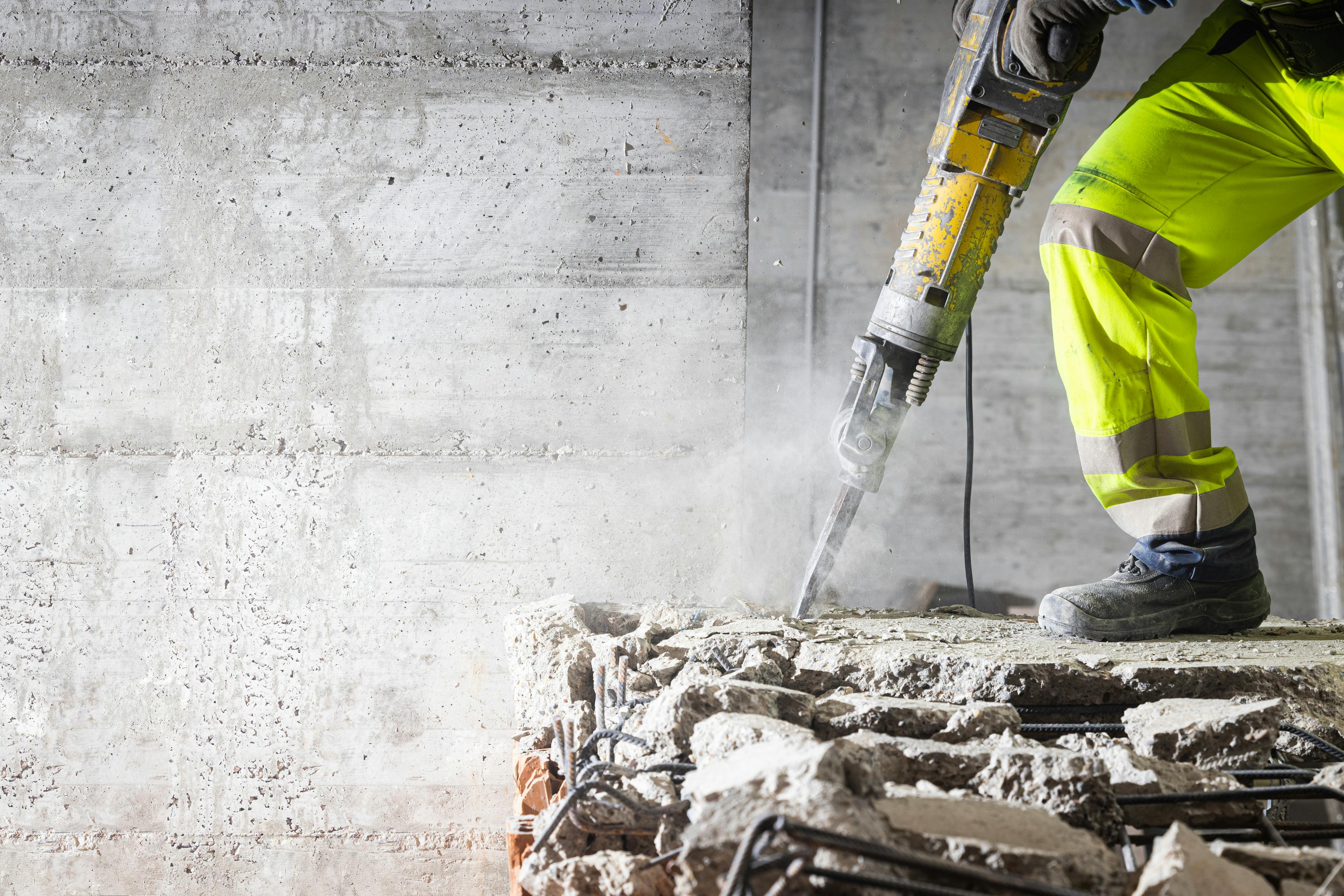 Das Bild zeigt einen Arbeiter in gelber Schutzkleidung, der mit einem Presslufthammer auf einer Baustelle arbeitet.