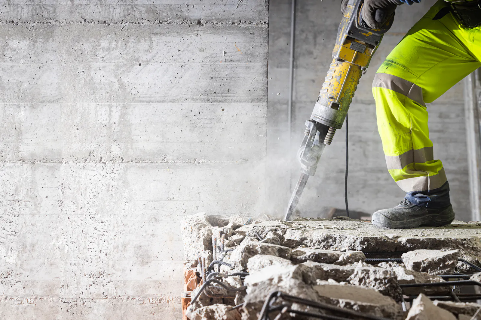 Das Bild zeigt einen Arbeiter in gelber Schutzkleidung, der mit einem Presslufthammer auf einer Baustelle arbeitet.